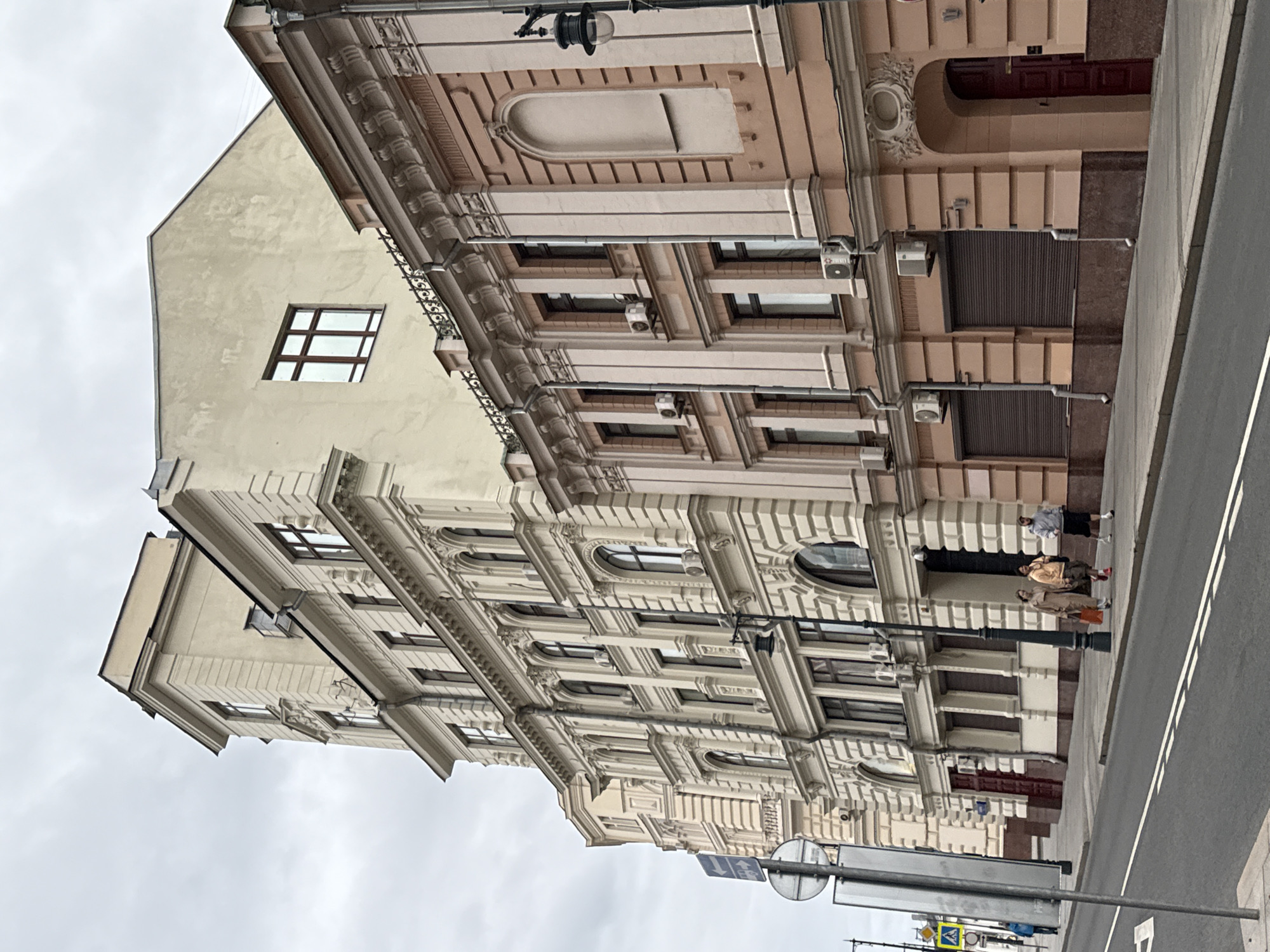 The image depicts a street view in Moscow, Russia, featuring a row of multi-story buildings with intricate architectural details. The buildings exhibit a classical style, with prominent cornices, decorative window frames, and arched entrances. The facade colors range from light beige to pale pink, with some darker accents. Several windows are visible, some with visible air conditioning units installed below them. A street lamp with a classic design stands on the sidewalk. In the foreground, a paved road with white lane markings runs parallel to the sidewalk. Three individuals are walking on the sidewalk, two women dressed in casual attire and one man wearing athletic wear. A blue pedestrian crossing sign and a circular traffic sign are mounted on a metal pole on the left side of the image. The sky is overcast and a muted grey, suggesting a cloudy day. The overall scene conveys a sense of urban architecture and daily life in a Moscow street.