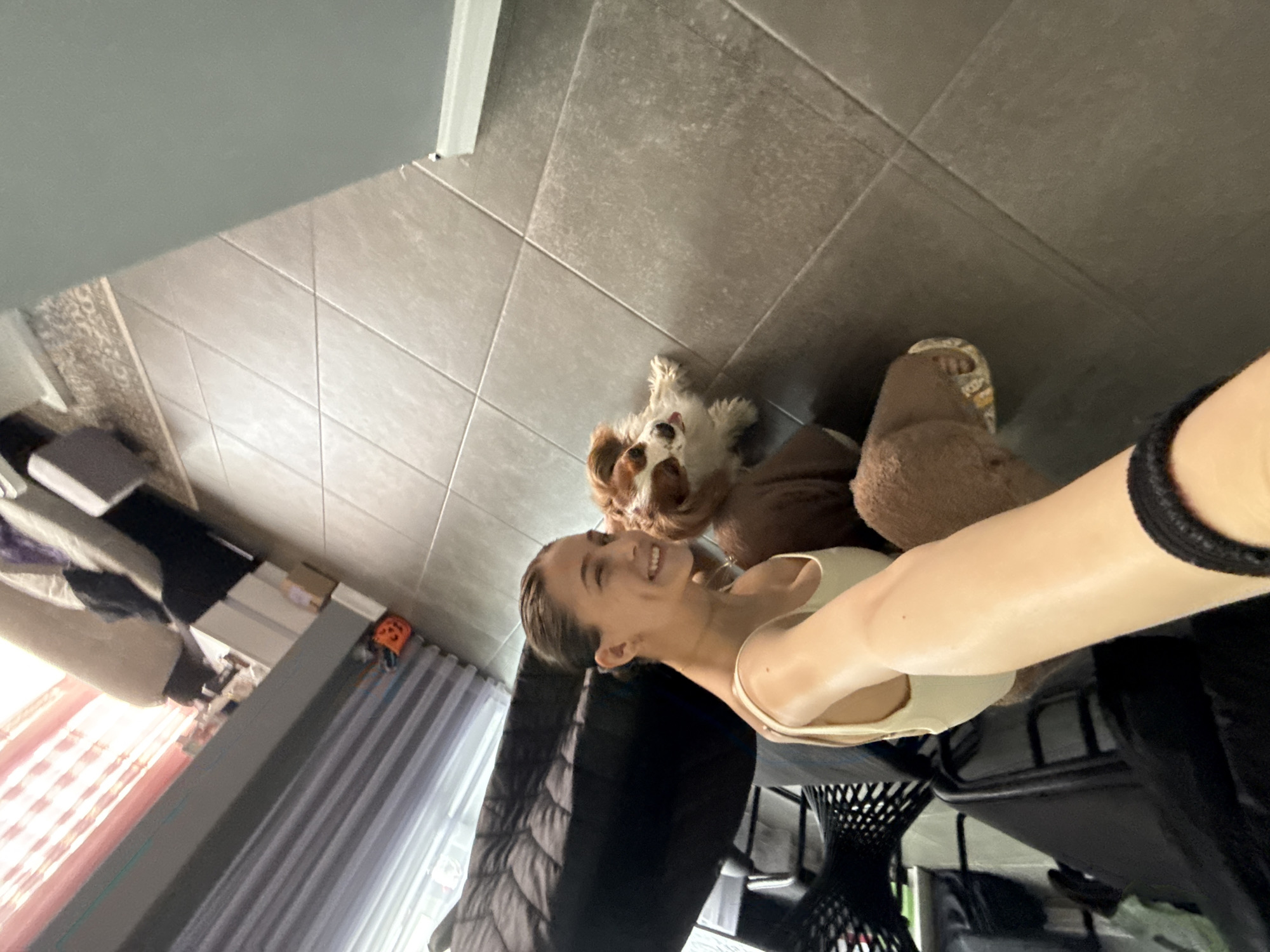 A woman is taking a selfie with her dog in an interior setting. The dog is lying on the floor with grey tiles, while the woman is standing with one foot in a brown slipper. In the background, there are curtains and what appears to be a bed or couch and a chair. Given the presence of Cyrillic text and the general style, this location is potentially within Sofia, Bulgaria.
