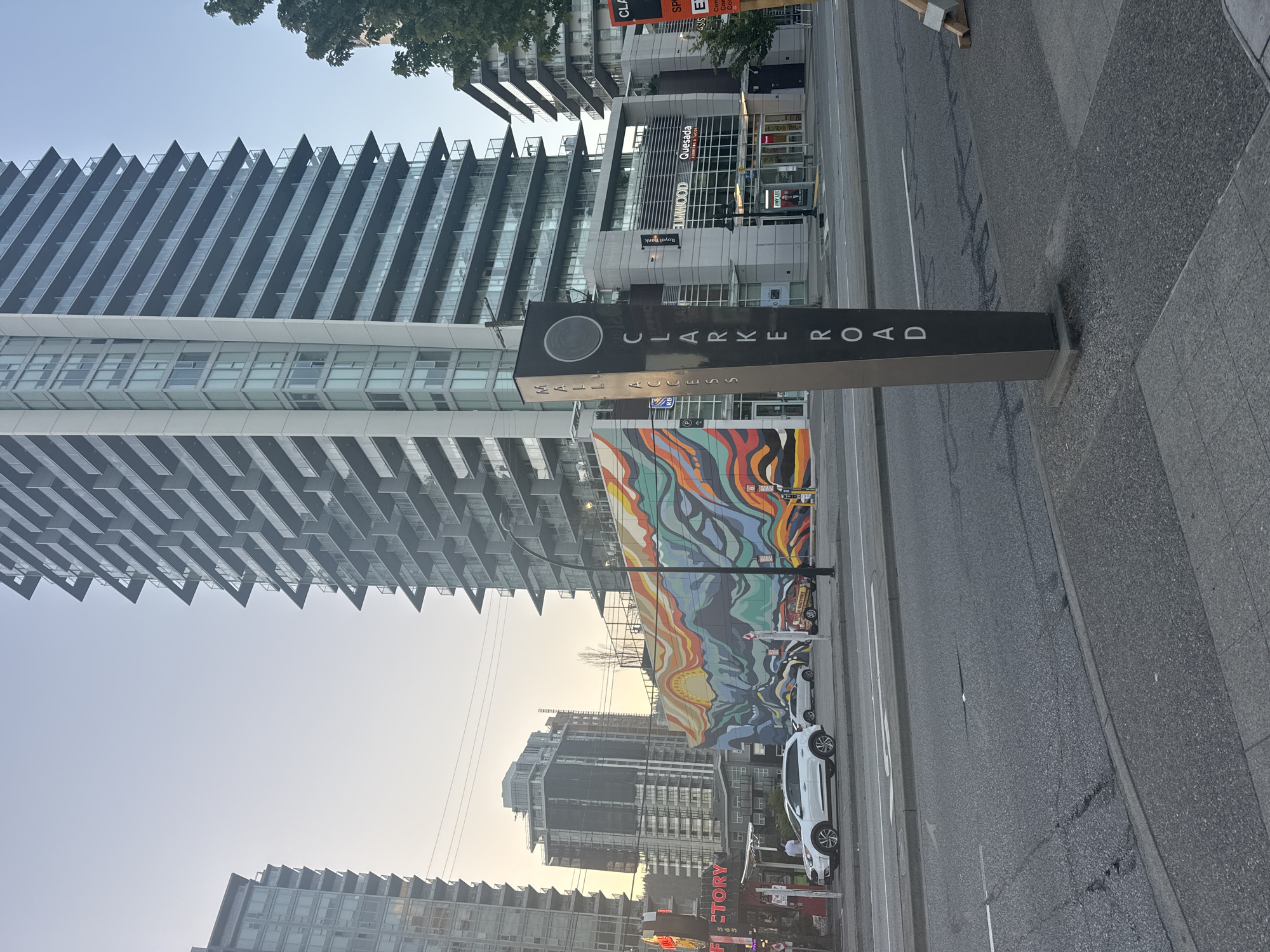 The image depicts an urban street scene, likely in Coquitlam, Canada, based on visible signage. A prominent pylon in the foreground displays "MAIL ACCESS CLARKE ROAD" vertically. Behind it, a large, colorful mural with abstract wavy patterns and a sun-like motif adorns the side of a building. Several high-rise residential buildings with balconies dominate the background, indicative of a dense urban environment. A white SUV and other vehicles are visible on the street. In the middle ground, commercial storefronts with names like "ROYAL BANK", "ELMWOOD", and "QUESADA" are present. The sky is clear and shows the soft light of early morning or late afternoon. The street is paved, with sidewalks and pedestrian crossings. Several trees are also scattered throughout the scene.