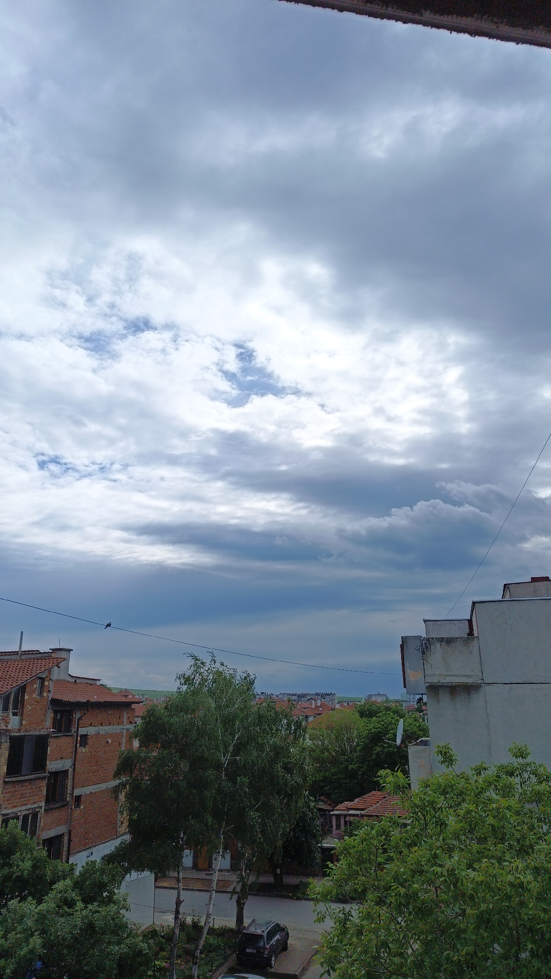 The image shows a cloudy sky over a cityscape. The buildings are mostly residential, with a mix of brick and concrete construction. There are trees and other vegetation visible in the foreground and middle ground, suggesting the scene is located in a residential area. A car is parked on the street. A bird sits on a wire. The location is a city of Unknown, Unknown.