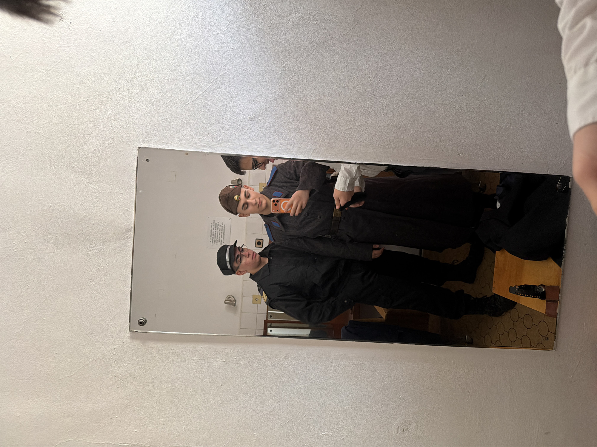A mirror reflects two people in what appears to be a bathroom. One person, wearing a brown cap with a red star emblem and a gray uniform, is taking a selfie with an orange cell phone. Another person, wearing a black cap, glasses, and a black uniform, stands nearby. The background includes a sink, a toilet paper dispenser, and a wooden cabinet. The reflection on the floor shows a pair of black shoes and what looks like a black case on a patterned floor. There is no information available to conclude the location in the city of Panagyurishte, Bulgaria.
