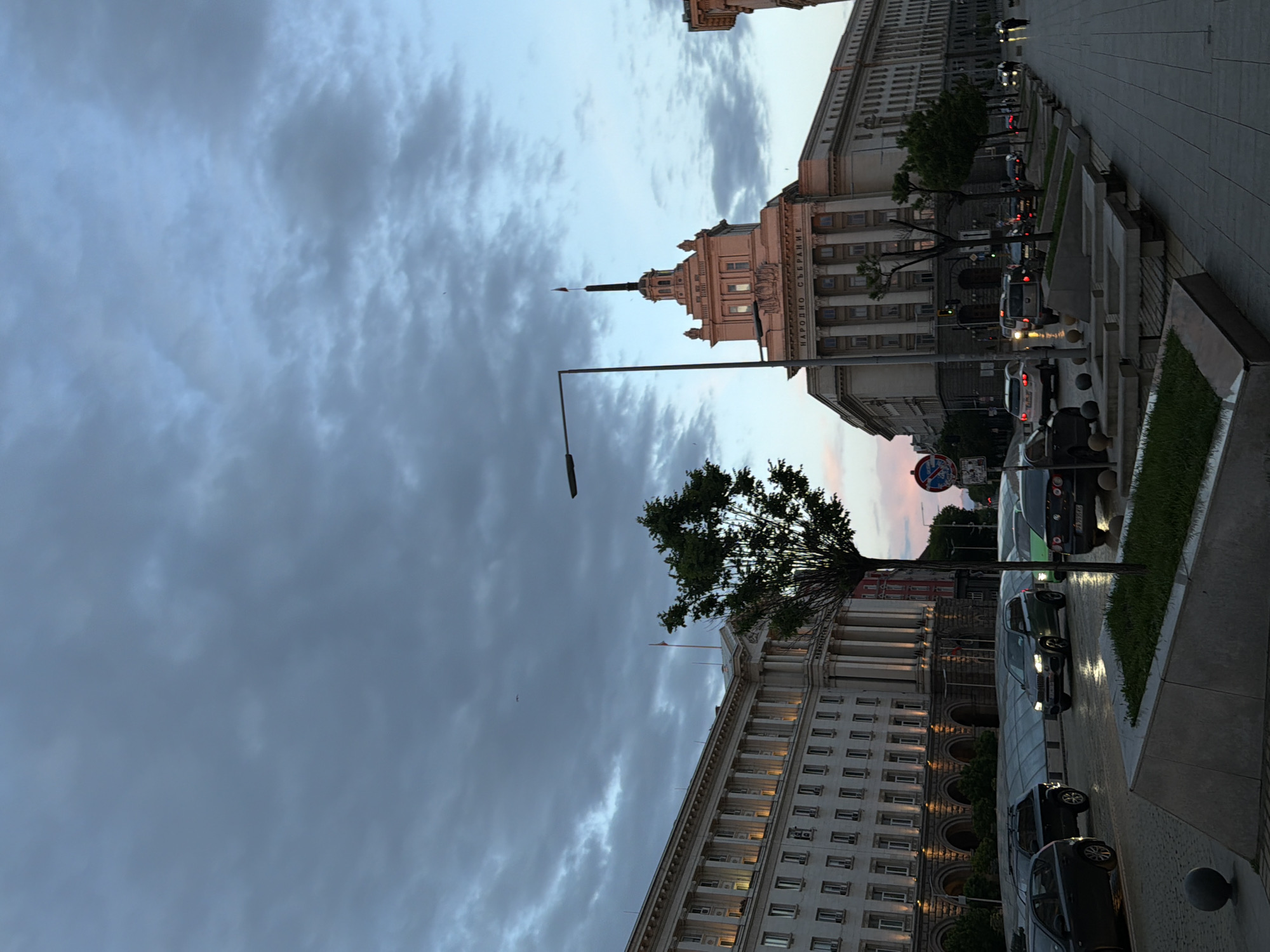 The image depicts a street scene in Sofia, Bulgaria, at dusk. The architectural elements are prominent, with the National Assembly building and other government buildings lining the street. Cars are parked along the road. Street lighting is visible, and the sky is overcast with dramatic clouds. There are trees in the background. The sidewalks are paved. There is no sign of people.