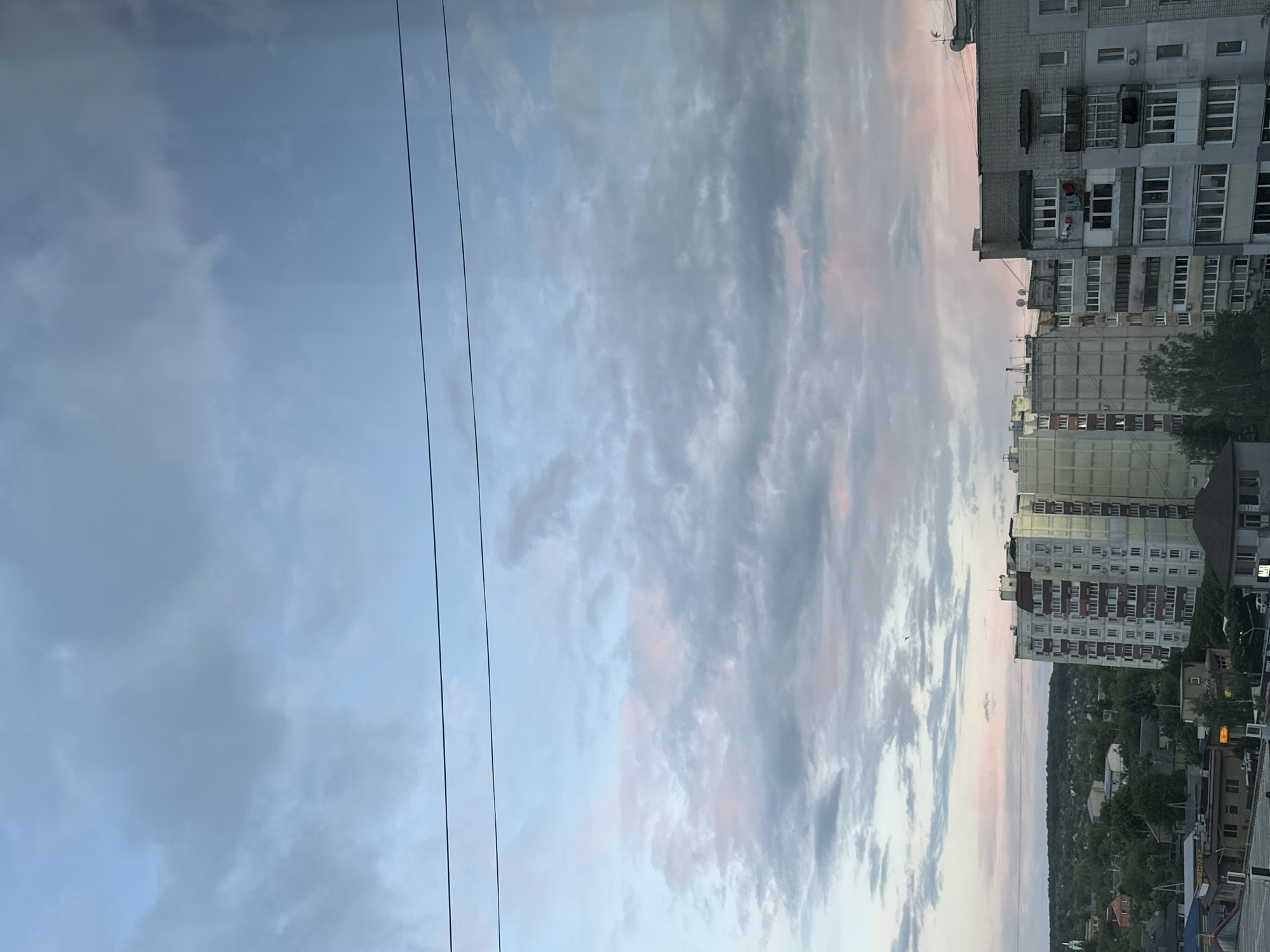 The image shows an urban landscape during twilight. In the foreground, two power lines stretch vertically across the left side of the frame, juxtaposed against a cloudy sky with hues of blue, gray, and pink. To the right, several multi-story apartment buildings dominate the skyline. The architectural style suggests post-Soviet housing with repetitive window patterns. Below the buildings, a scattering of trees and rooftops indicates a mix of residential and commercial areas. A streetlamp is visible near the bottom edge. The scene suggests a location within a residential district near the city center, characterized by aging infrastructure and dense housing.