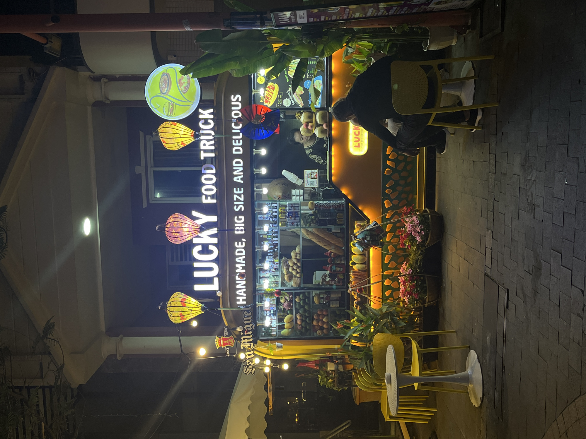 The image shows a brightly lit food truck called "Lucky Food Truck" located in Đà Nẵng, Vietnam. The truck has an orange and yellow color scheme with tropical plants adorning the top and sides. It is stocked with various food items, including baguettes, fruits, and drinks. A person is standing inside the truck preparing an order. A person is seated on a yellow chair eating or observing. The area in front of the truck is arranged with yellow chairs and a tall standing fan. String lights are hung above the seating area. The signage includes the name of the truck, "Lucky Food Truck," with the description "Handmade, Big Size and Delicious" and a San Miguel beer logo. A sign on the truck lists menu items and prices, including Banh mi and sandwiches. The setting is at night on a paved street.