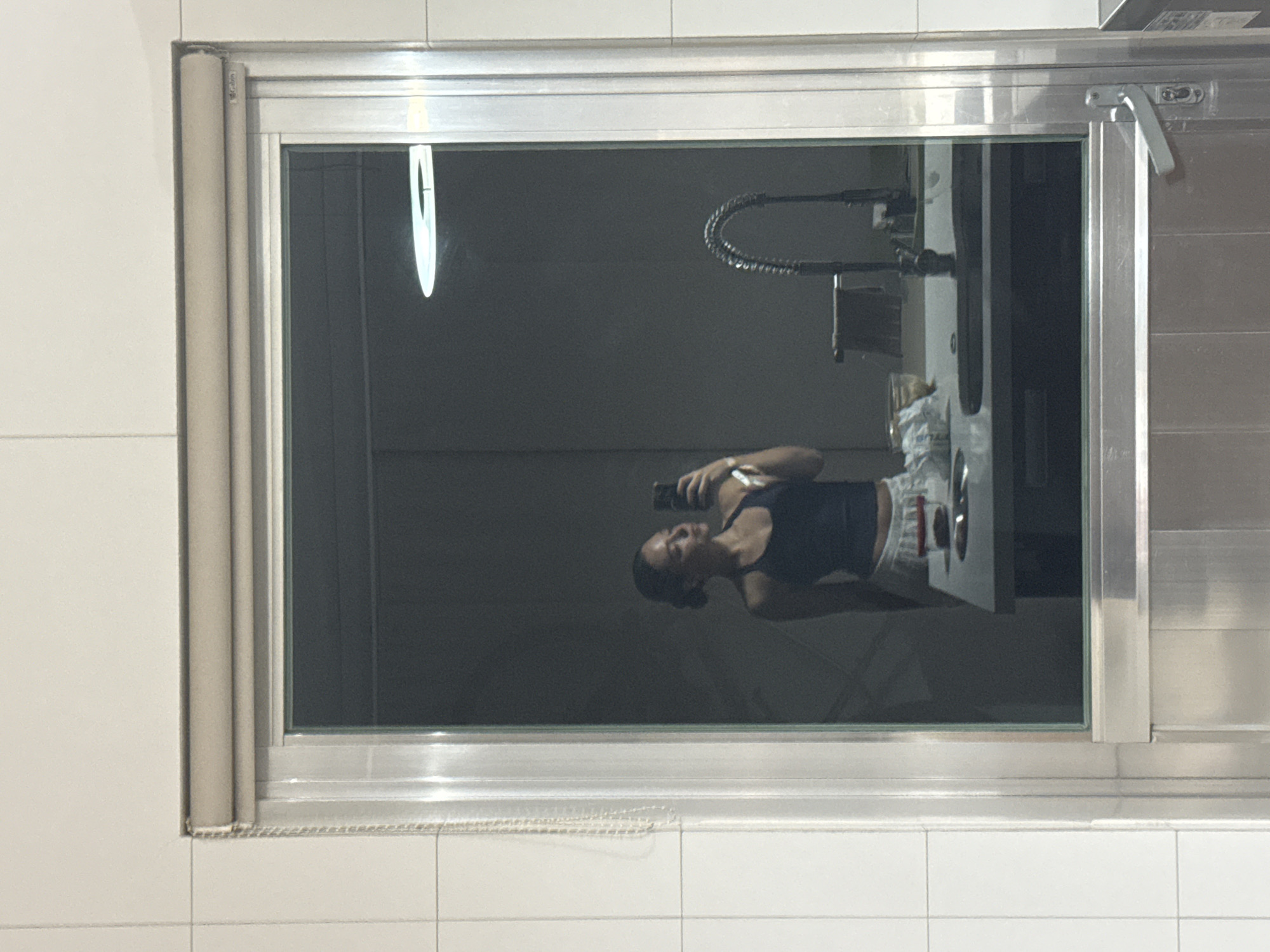 The image depicts a reflection in a window or mirrored surface of a person holding a smartphone, likely taking a photo or video. The reflected environment suggests a kitchen or food preparation area, as there's a sink with a flexible hose faucet and metallic surfaces. The surrounding area appears to be tiled, with white tiles visible on the wall around the window or mirror. The reflection captures a person in a tank top and shorts. There is no information in the image that indicates the specific location of San Bernardino Tlaxcalancingo, Mexico.