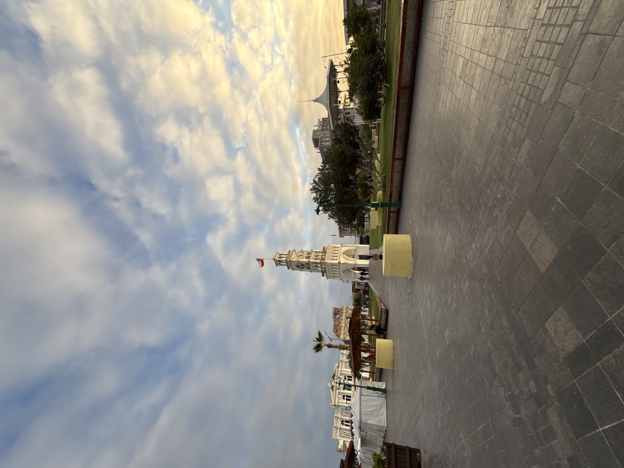 The image depicts the Plaza Prat in Iquique, Chile, at dusk. A tall, white clock tower, topped with a Chilean flag, stands prominently in the center of the frame. The sky is filled with a dramatic mix of white and golden clouds, illuminated by the setting sun. In the foreground, a paved plaza with intricate stone patterns reflects the soft light. To the left, white tents suggest a market or event setup. Several people are visible, some walking near the clock tower and others seated under a covered pavilion. Lush green trees and a well-maintained lawn flank the plaza, with other buildings visible in the background, including a grand white structure to the left and a modern building with a clock face to the right. The overall atmosphere is serene, with the warm glow of the sunset casting long shadows across the scene.
