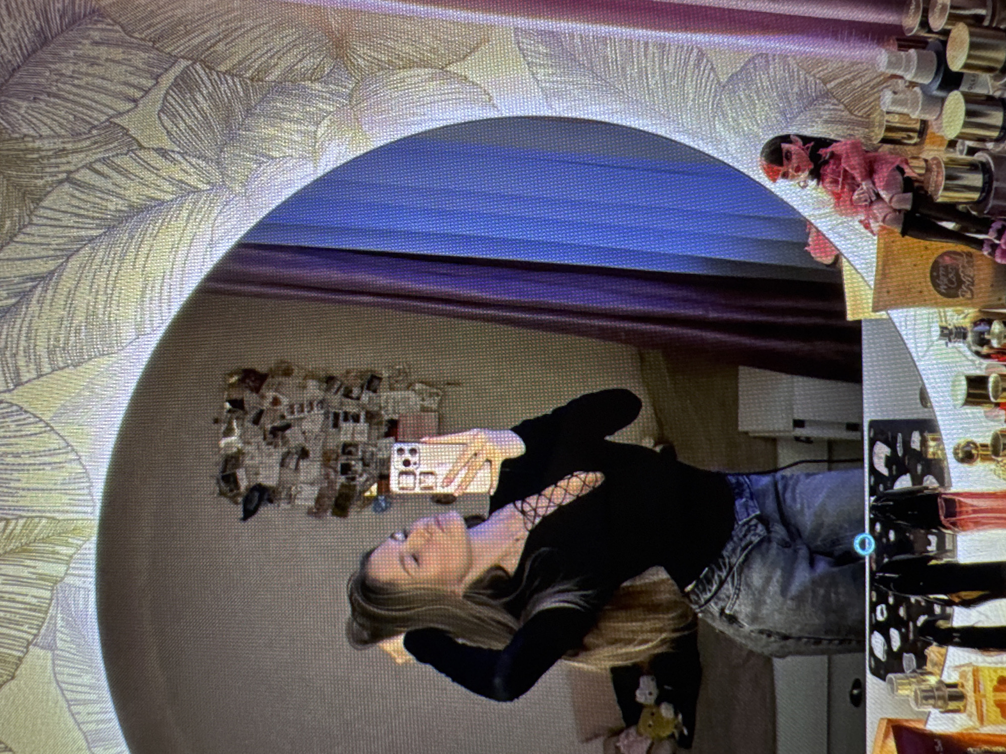 A young woman with long blonde hair is taking a selfie in a large, oval mirror. She is wearing a black long-sleeved top with a lace-up neckline and grey jeans. Her hair is pulled back with one hand, and she is holding her phone in the other, angled to capture her reflection. The mirror is framed by a decorative wallpaper with a leaf pattern. In the reflection, a purple curtain is visible behind her. In the foreground, on a shelf below the mirror, are several bottles of perfume and a doll. The background behind the woman shows a bed and a wall decorated with many small photographs.