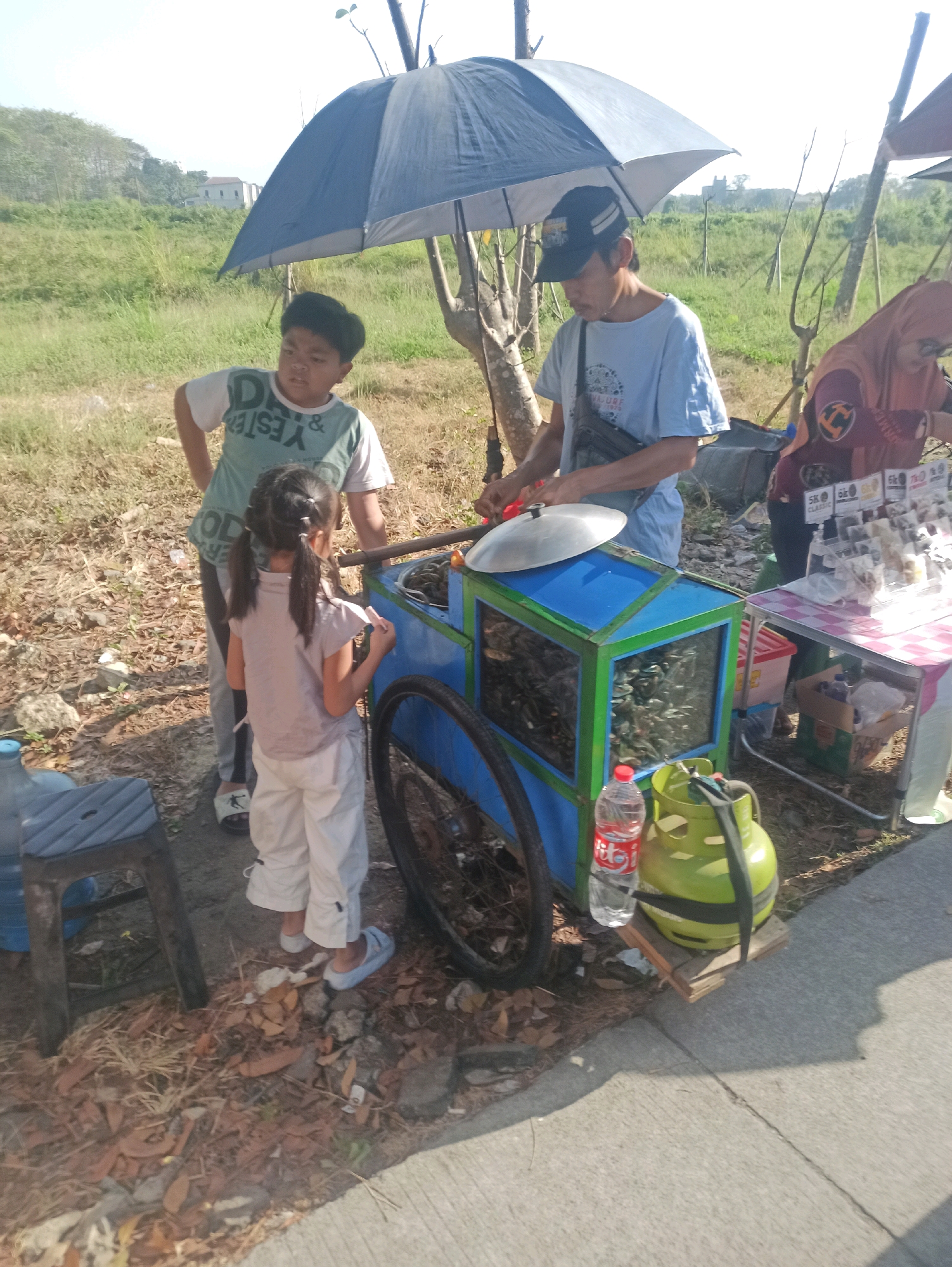 The image depicts an outdoor scene with a food cart being operated by an adult male, who is wearing a blue t-shirt and a dark baseball cap. He is standing next to a blue and green food cart, which is equipped with a metal lid on a pan. A young boy, wearing a green and white t-shirt, stands to the left of the cart, and a young girl, in a light-colored shirt and white pants, stands in front of the cart holding a small item. A woman, wearing a hijab and an orange and purple top, is visible behind a table to the right, where various items are displayed. The food cart has a glass display case filled with what appear to be food items. A green propane tank and a bottle of water are situated on the ground next to the cart. The environment appears to be a grassy area with some dry leaves on the ground, and trees in the background under a bright sky. The scene is set on the side of a paved road, indicating a likely location in Telagamurni, Indonesia, where such street-side vendors are common.