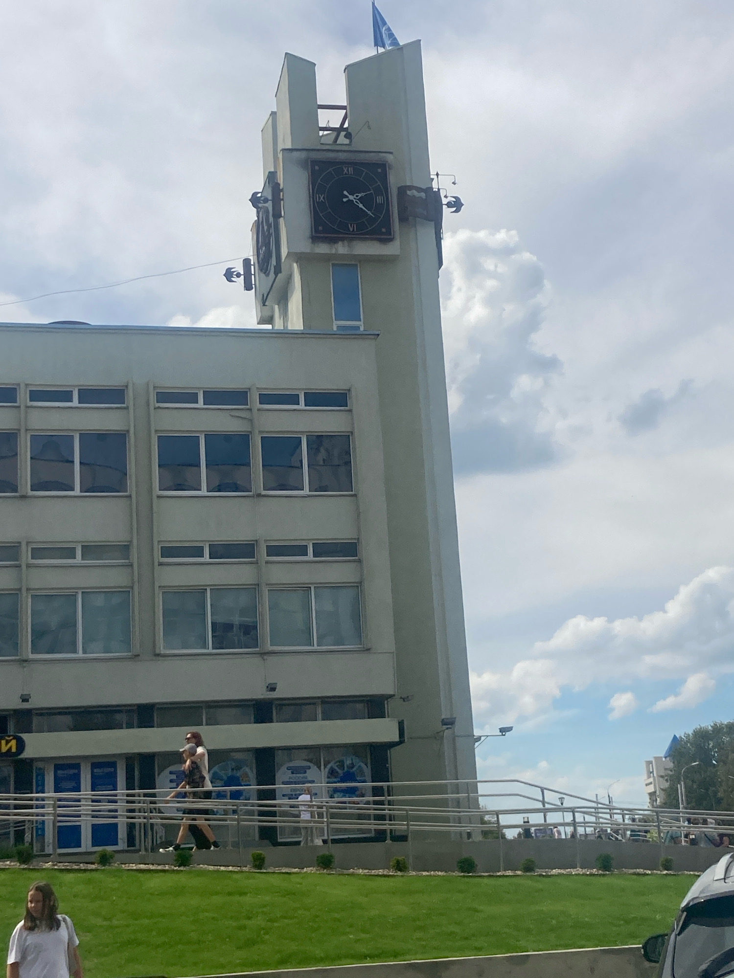 The image shows a tall building with a clock tower, located in the city of Brest, Belarus. The clock tower has a large clock face with Roman numerals and intricate metal bird sculptures attached to its side. The building itself is light-colored, with multiple stories featuring rows of rectangular windows. In front of the building, there is a green lawn with a few bushes. A few people are visible in the scene, including a woman walking in the foreground and a couple walking on a ramp in front of the building. The sky is partly cloudy, with a blue sky visible between white clouds.