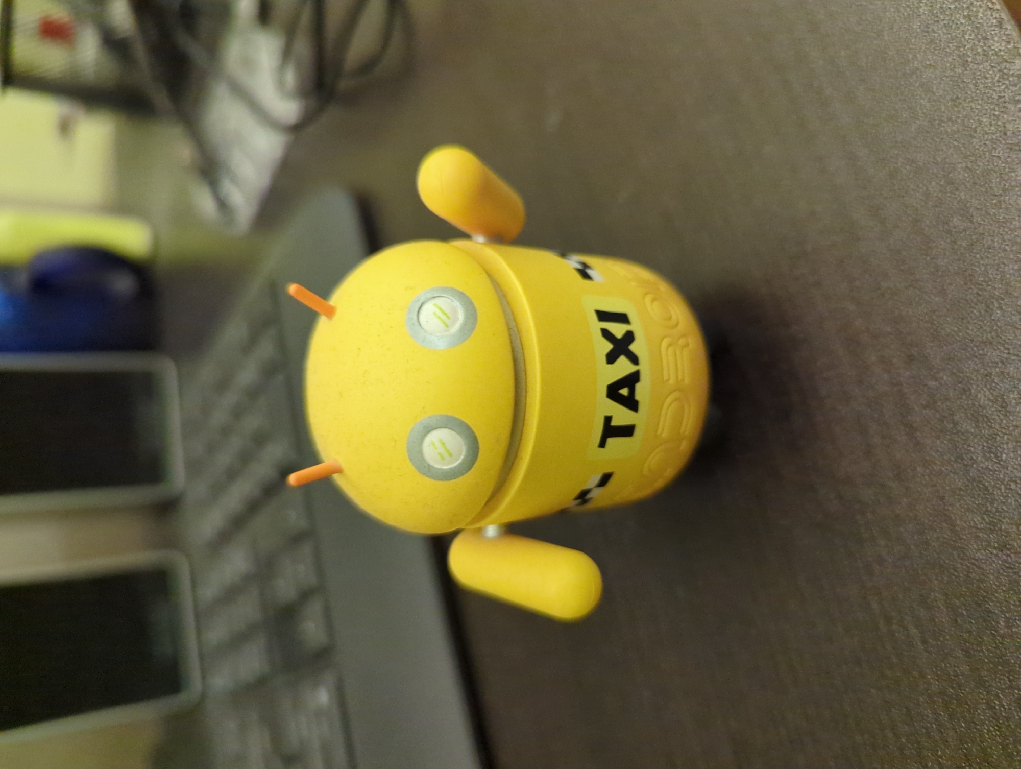 The image shows a yellow, cylindrical Android robot toy that is labeled "TAXI". It is placed on a dark surface, likely a table or desk, next to a computer keyboard. The robot toy has two yellow arms, two orange antennae, and two silver "eyes" on its face. The background is blurry but suggests an indoor environment with some office equipment or other objects. The location is not explicitly mentioned in the image or implied through the visual content. Therefore, it is impossible to ascertain whether the location is Minsk, Belarus.