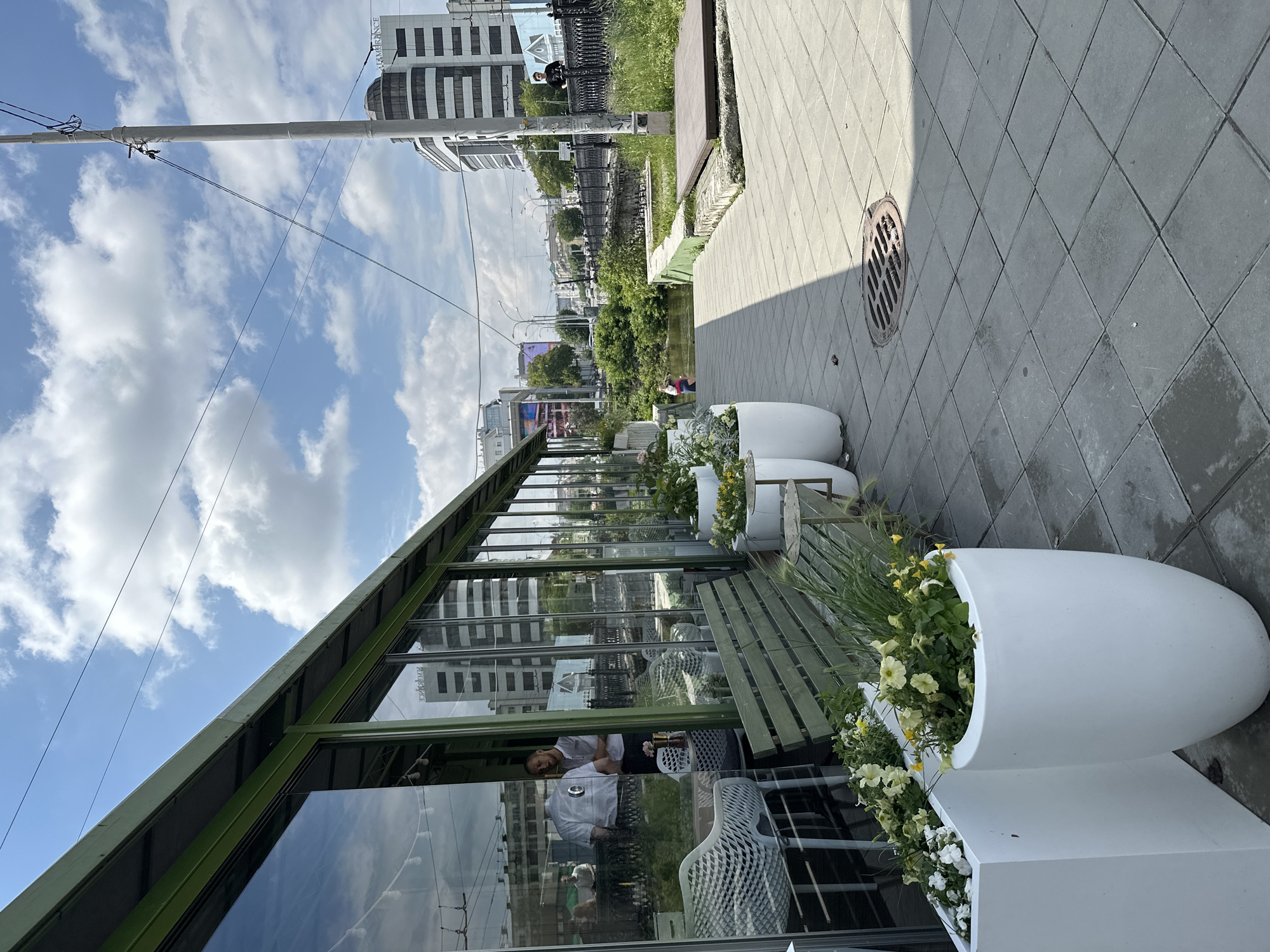 The image depicts an outdoor seating area of a cafe or restaurant in Yekaterinburg, Russia, situated along a waterway. The building features a green-trimmed facade with large glass windows, reflecting the cloudy sky and surrounding urban landscape. Several white planters containing yellow and white flowers and greenery are placed along the sidewalk in front of the establishment. Green wooden benches are also visible.
Inside the cafe, reflections in the glass reveal a few patrons and staff. Through the windows, a waterway can be seen, spanned by a bridge with an ornate black metal railing. Beyond the waterway, modern buildings, including a prominent multi-story structure with a curved facade and striped detailing, are visible. A street with traffic and streetlights runs parallel to the waterway.
On the sidewalk in the foreground, a metal manhole cover is set into the grey, tiled paving stones. A lamppost stands on the right side of the image. A person in a dark t-shirt is standing on the bridge railing. Another person, wearing a red top and dark bottoms, is visible near the waterway. The overall impression is an urban scene on a bright, cloudy day, with elements of nature (the waterway and plants) integrated into the cityscape.