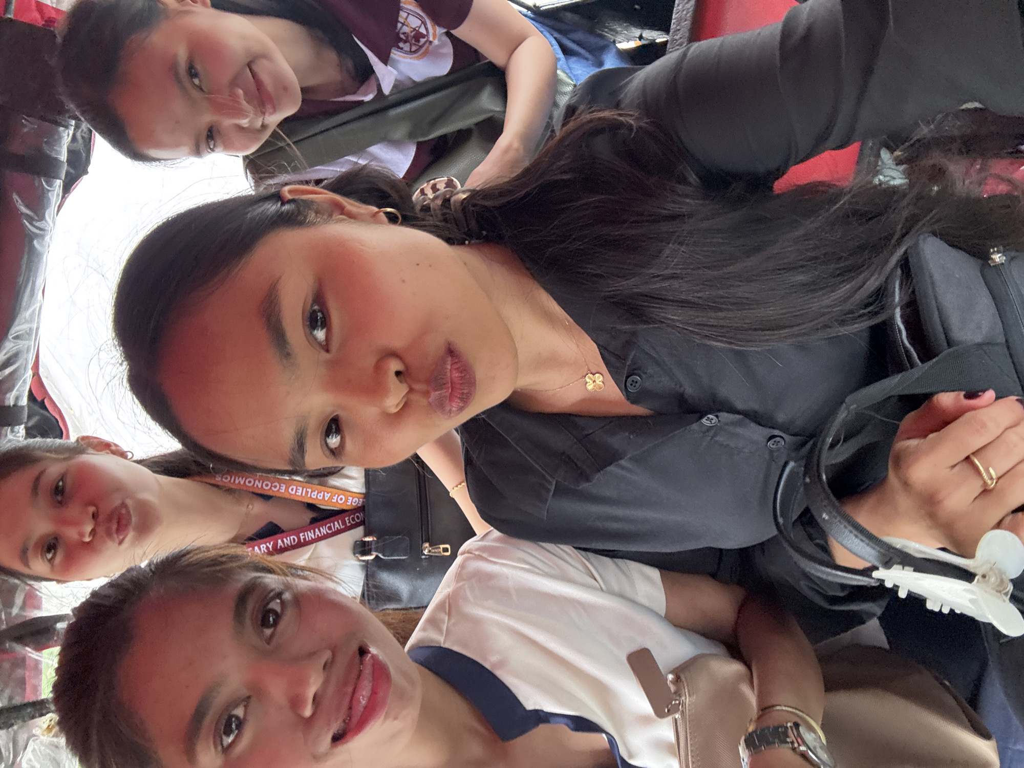 Four young women are posing for a selfie in what appears to be the interior of a vehicle, likely a tricycle common in Davao City, Philippines, given the context. The women are seated closely together. The woman in the foreground on the right is wearing a black collared shirt and has her hair pulled back. She is making a "duck face" and wearing a necklace with a clover-shaped pendant and a gold ring on her right hand. To her left is another woman in a white collared shirt with navy blue trim, also making a "duck face" and sticking her tongue out slightly. She is wearing a watch on her left wrist and holding a light brown purse. In the background, two other women are visible. One is wearing a white shirt with a lanyard around her neck that reads "COLLEGE OF APPLIED ECONOMICS," and she is also making a "duck face." The woman on the far right is wearing a maroon and white collared shirt with a school emblem. The interior of the vehicle shows a red upholstered seat and some reflective white material, possibly the roof. The lighting suggests it is daytime.