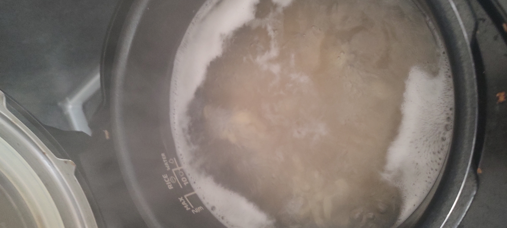 A top-down view of a rice cooker filled with boiling water and rice. The water is cloudy and has a foamy layer around the edges. The rice cooker is black and has markings indicating the water level for rice. The location is Unknown, Unknown.