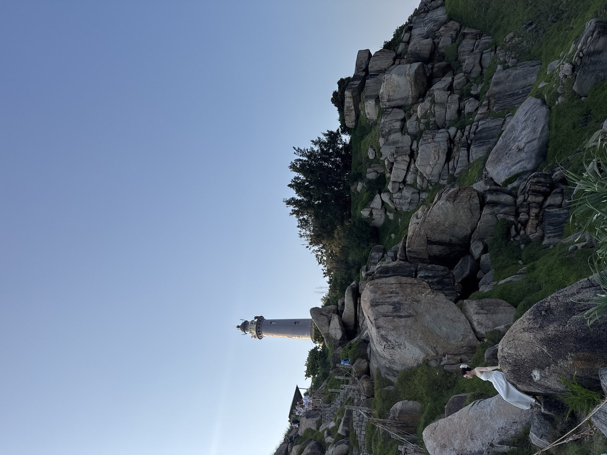A sideways, long shot captures a lighthouse, rocks, vegetation, and a person. The lighthouse, a tall structure with a cylindrical shape, stands against a clear, light blue sky. Positioned on a rocky slope, it is flanked by a dense cluster of grey rocks, some of which are interspersed with patches of green vegetation. A few trees dot the area around the lighthouse. A wooden staircase leads towards the structure, with several people visible on the lower steps. A person dressed in a white garment is seen on the right side of the image, perched on a large rock. The location is listed as Unknown, Unknown.