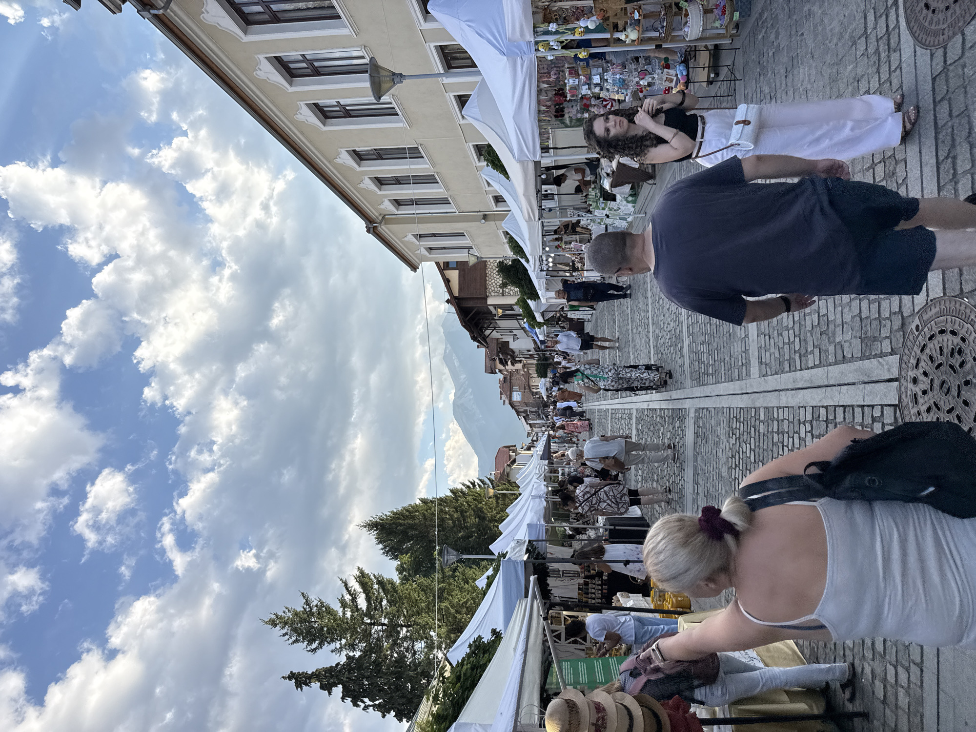 The image depicts a vibrant street market in Bansko, Bulgaria, with numerous stalls set up along a cobblestone street. The market is bustling with activity, as various individuals browse the merchandise. The stalls are covered with white awnings, creating a shaded pathway for shoppers. The buildings lining the street are traditional in style, with ornate facades and large windows. In the background, majestic mountains rise against a partly cloudy sky, adding to the scenic beauty of the location. The atmosphere appears lively and engaging, suggesting a popular community event or a typical market day in Bansko.