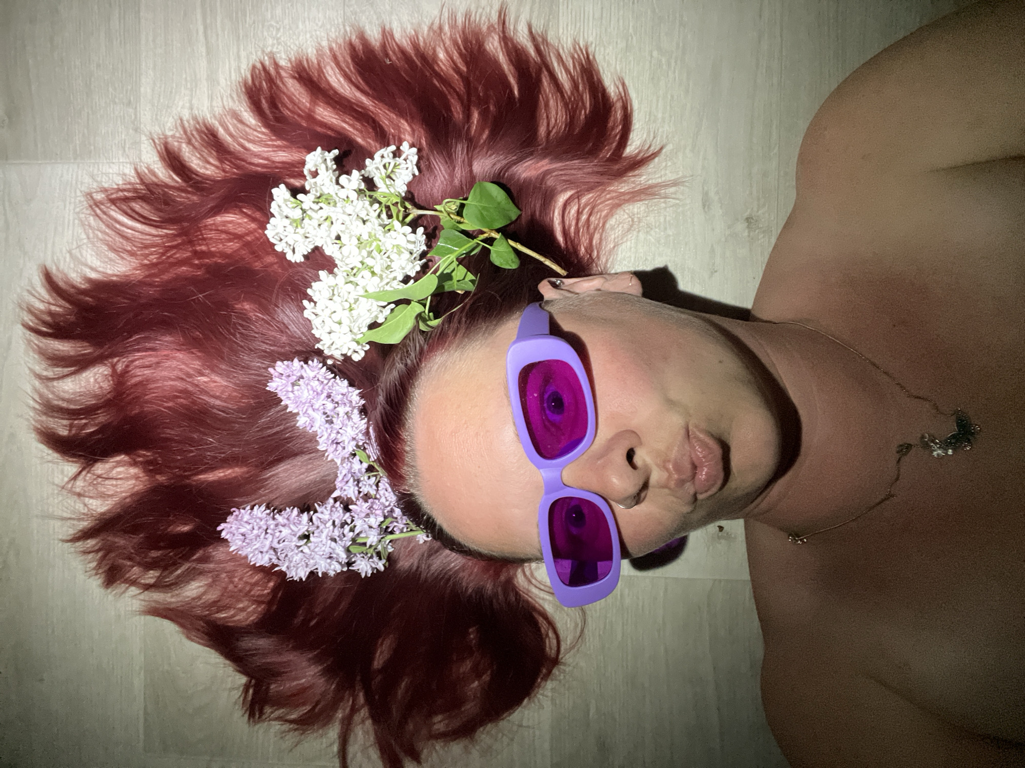 A woman is lying on a floor. She has red hair adorned with white and purple flowers, and she is wearing purple sunglasses. A gold necklace is visible on her chest. The floor appears to be made of light-colored wood. The image does not contain enough information to confirm that the location is in the city of Chaikovsky, Russia.
