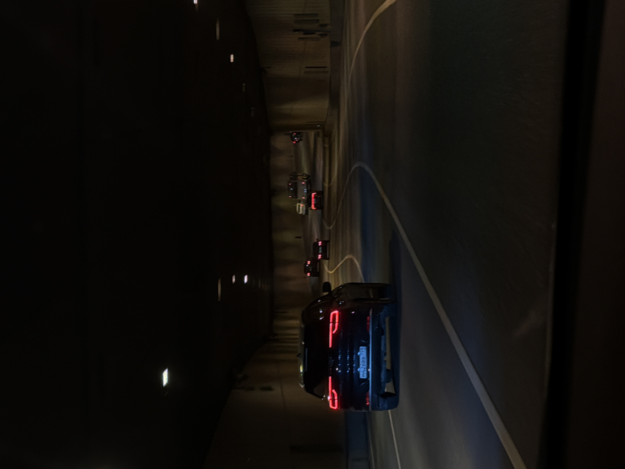 This is a low-light, horizontal, outdoor photograph taken at night from inside a vehicle traveling through a tunnel in Moscow, Russia. The tunnel is a concrete structure with overhead lighting. In the foreground, a dark-colored SUV with its red taillights illuminated is driving in the right lane. The SUV has a license plate that appears to read "M426BB 775". Other vehicles are visible further ahead in the tunnel, including a bus and several cars, all with their lights on. The road surface is dark asphalt with white lane markings. The tunnel walls are visible on both sides of the roadway.