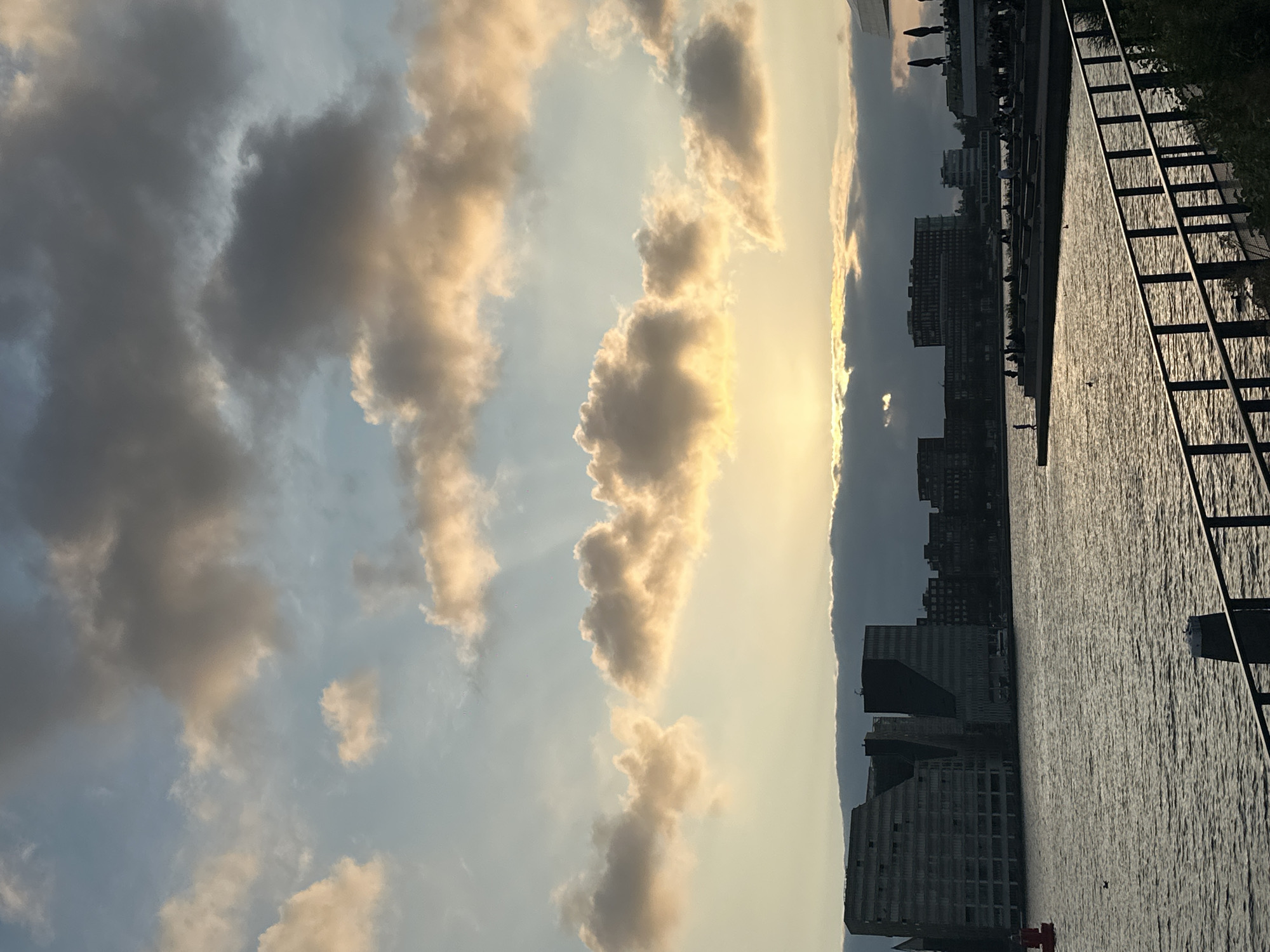 This image captures a scenic view of the Amsterdam skyline at sunset, likely from the Oostenburg neighborhood. The sky is filled with a dramatic mix of light and dark clouds, with the sun peeking through, casting a warm glow on the scene. In the foreground, a calm body of water reflects the colors of the sky and the surrounding architecture. On the left side, a prominent red buoy is visible in the water. Across the water, a row of modern buildings forms the city's silhouette, with distinctive architectural styles. A promenade or pier extends into the water, populated by several individuals enjoying the evening. Some are seated on tiered steps, while others stroll along the waterfront. A dark metal railing runs along the bottom right of the image, suggesting the viewpoint is from a walkway or balcony. The overall atmosphere is serene, highlighting the interplay of natural light and urban landscape.