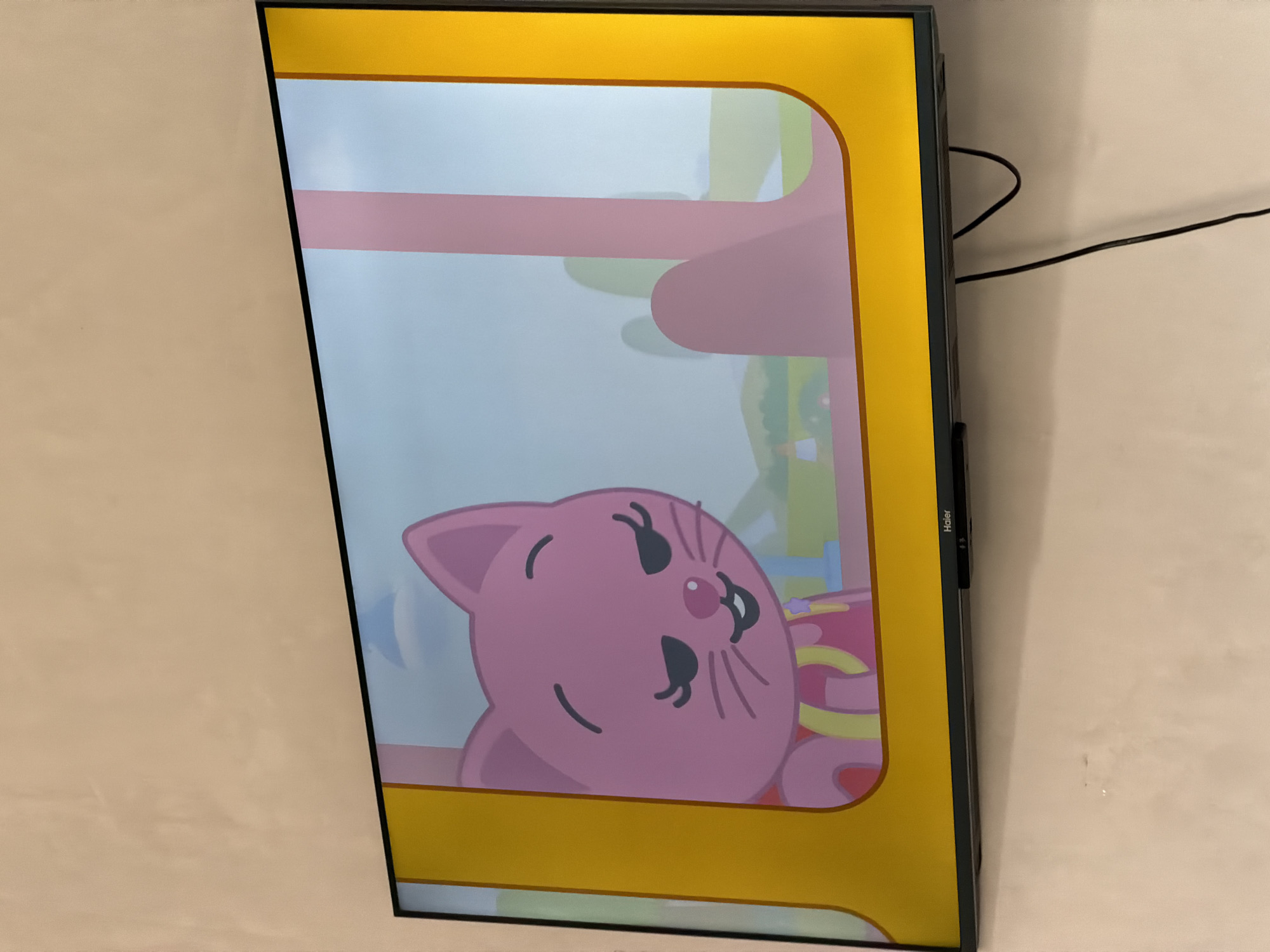 A flat-screen television mounted on a light-colored wall displays a cartoon image of a pink cat with closed eyes and a slight smile. The cat is wearing a pink outfit with yellow straps and a small star emblem. The background of the cartoon is a bright sky with some green hills. The TV itself is black with a thin black bezel. A black power cord hangs down from the bottom of the TV. The location is not identifiable as Krasnyi Krym, Russia.