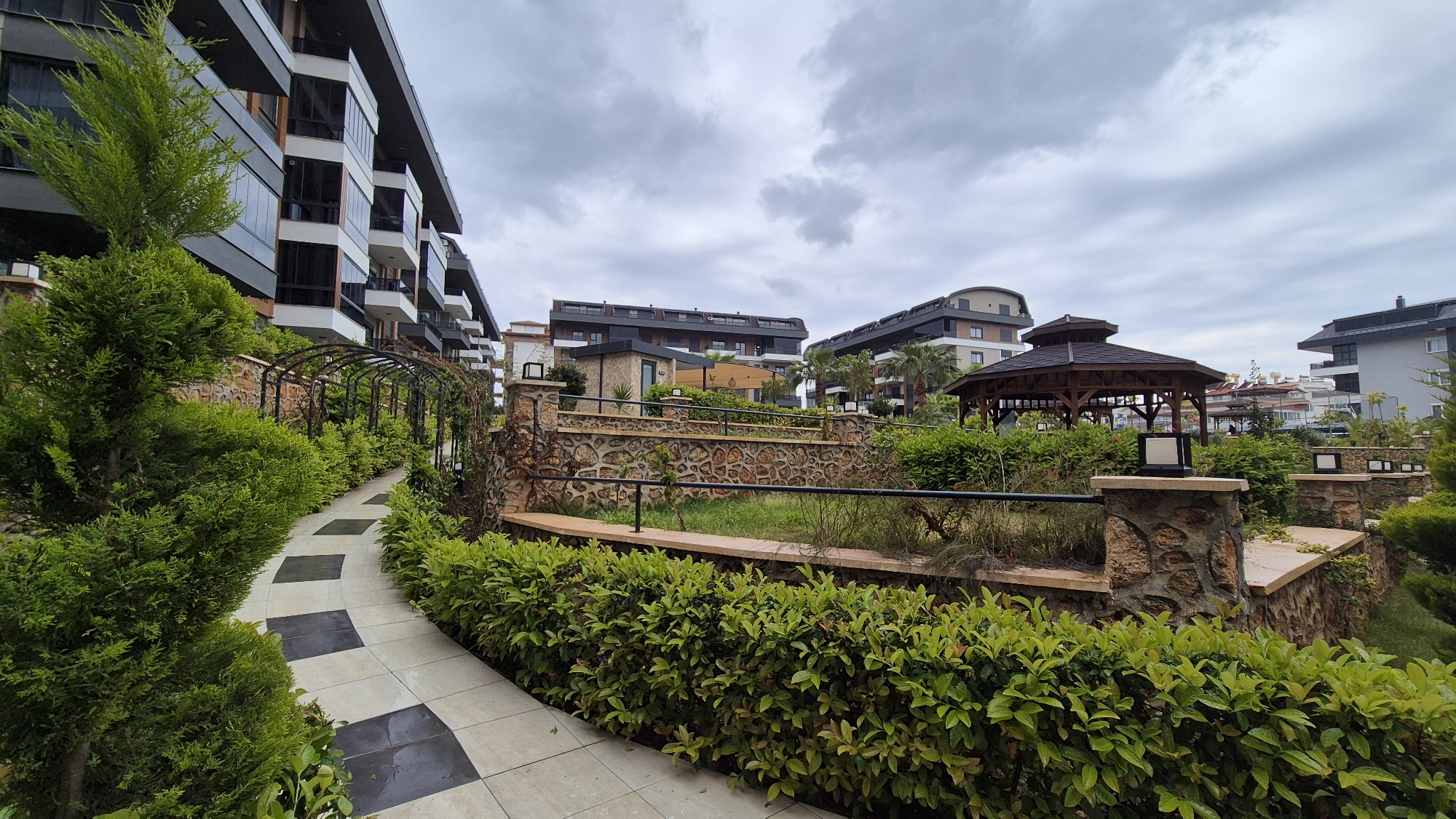 The image depicts a residential area in Alanya, Turkey, under a cloudy sky. The scene includes apartment buildings with modern architecture, featuring balconies and dark gray trim. A landscaped garden pathway with black and white checkered tiling winds through manicured hedges and low stone walls. A gazebo stands amidst the greenery, and decorative light fixtures are mounted on stone pillars along the walkway. The foreground is dominated by vibrant green foliage, while other apartment buildings and a variety of trees are seen in the background.