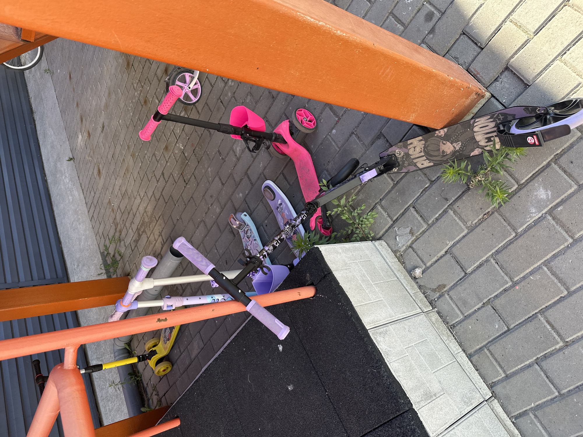 In Novoaltaysk, Russia, a collection of children's scooters are scattered on a paved area near a structure with orange and orange-red supports. A yellow scooter with black handlebars is positioned near a grey pipe. Adjacent to it are two smaller scooters, one white with light purple handlebars and accents, and another similar scooter, also with light purple handlebars. Further to the right, a pink three-wheeled scooter with a pink basket on the handlebars is visible. At the bottom of the image, a black scooter with purple accents and graphics, including anime-style artwork and text, is lying on the ground. The environment includes a grey tiled border along a black rubberized surface, and patchy green vegetation growing between the paving stones.