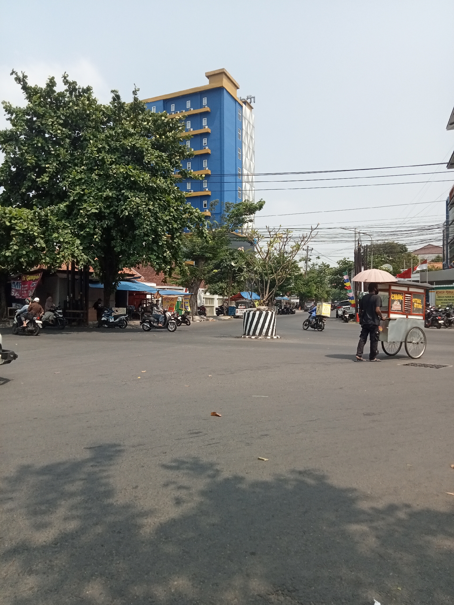 The image depicts a street scene in Semarang, Indonesia, with a focus on the intersection where a street vendor with a food cart is visible. The vendor, a man wearing a black shirt and pants, stands beside a cart with an umbrella overhead. The cart has signage that reads "CIROK" and "OTAK OTAK" in Indonesian, indicating food items for sale. The cart itself is mounted on two wheels and appears to be made of metal and wood.
In the background, a tall, modern blue building with yellow accents stands prominently, suggesting an urban environment. Several trees with dense green foliage are present, providing shade and breaking up the cityscape. The street is paved and shows signs of traffic, with several motorcycles parked or in motion. Some of these motorcycles have riders.
The environment appears to be a typical street in a city with commercial activity. The lighting suggests it is daytime, likely with clear skies. The scene captures a moment in the daily life of the city, highlighting the presence of street vendors and the flow of traffic.