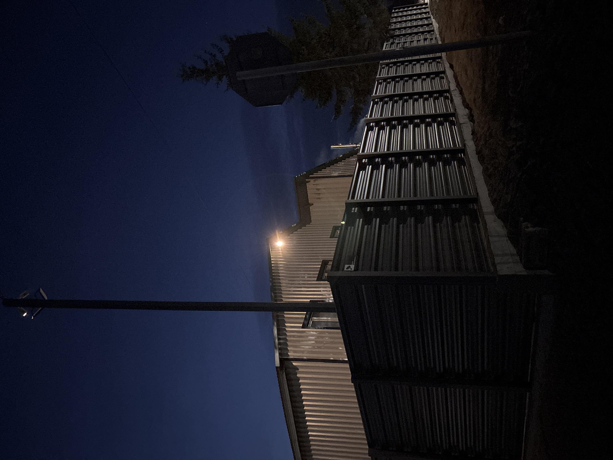 Low-angle night shot. A building with corrugated metal siding is illuminated by a single light fixture. A metal fence runs along the right side of the building. A wooden utility pole with a light fixture extends from the lower-left corner into the dark blue sky. Trees and a tower are visible in the upper-left corner. The right side of the image is in deep shadow. Location inferred to be Фоки, Russia due to query context.