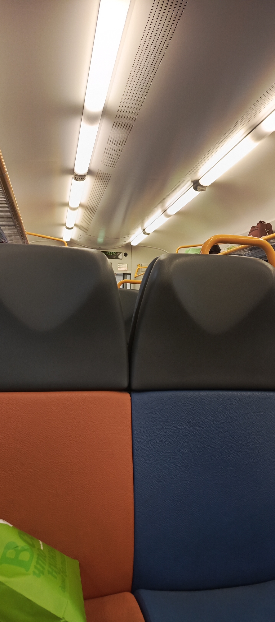 The image shows the interior of a train carriage. The focus is on the seating area, with two rows of seats in the foreground. The seats have dark gray backs and orange and blue cushioned bases. Above the seats, the ceiling of the carriage is visible, lined with fluorescent lights. Some of the lights appear to be flickering or reflecting unevenly. In the background, a yellow handrail is visible, as well as luggage on an overhead rack. There is a digital display showing "+22°C" and some text on a white sign. A green bag with some text is partially visible in the lower left corner of the frame. The scene suggests the train is in motion or stopped at a station. The lighting indicates it is likely daytime or within a well-lit interior. The presence of luggage and seating suggests travel.