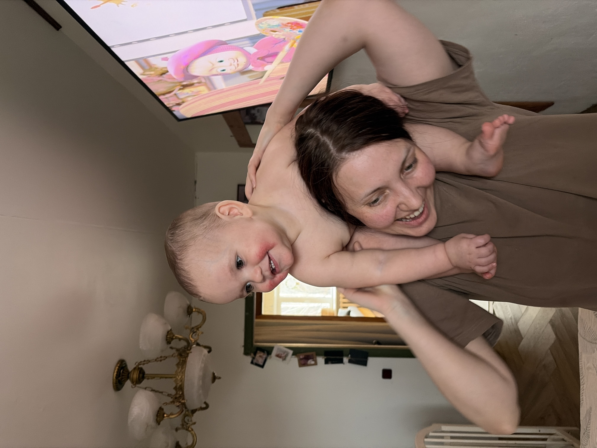In a residential interior located in Chaikovsky, Russia, a woman with dark hair, wearing a brown t-shirt, is depicted from the chest up, smiling and looking towards the lower right. She is carrying a fair-skinned infant on her shoulders. The infant, unclothed from the waist up, exhibits red marks on both cheeks and is smiling while looking towards the upper left. The woman's right arm is extended upwards, supporting the infant's left leg, while the infant's left hand rests on the woman's head.
The background shows a light-colored wall and ceiling. A multi-arm brass-toned chandelier with five frosted glass shades is mounted on the ceiling in the upper left quadrant. To the right, a large, dark-framed flat-screen television displays an animated cartoon character resembling "Masha" from "Masha and the Bear," wearing a pink beret and holding an artist's palette and brush. A doorway with dark trim is visible behind the subjects, with several framed photographs vertically arranged on the wall beside it. Through the doorway, another room with a window is partially visible. A white wooden crib is partially visible in the lower left corner. The flooring in the foreground and through the doorway appears to be light-colored wood or laminate laid in a herringbone pattern.