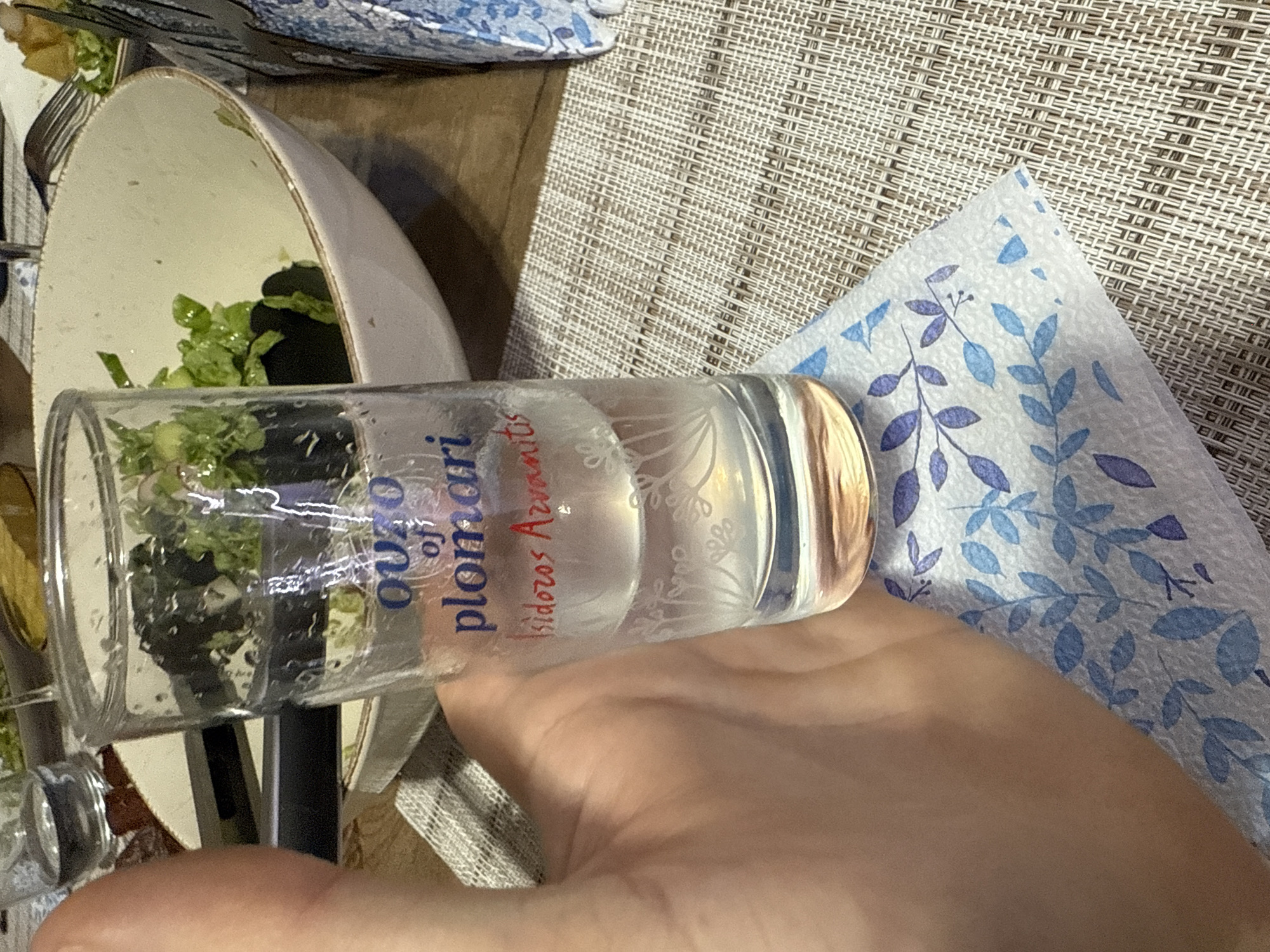 A horizontal, close-up shot shows a glass filled with liquid, likely ouzo, held in a person's hand. The glass, labeled "Ouzo of Plomari Isidoros Arvanitis," is decorated with a subtle floral design. Behind the glass, a partially eaten salad sits in a white bowl with a tan rim. A fork rests on the edge of the bowl. A beige woven placemat is visible beneath the bowl and the person's hand. A blue and white patterned napkin is adjacent to the hand. There is no indication of Sofia, Bulgaria, in the image.