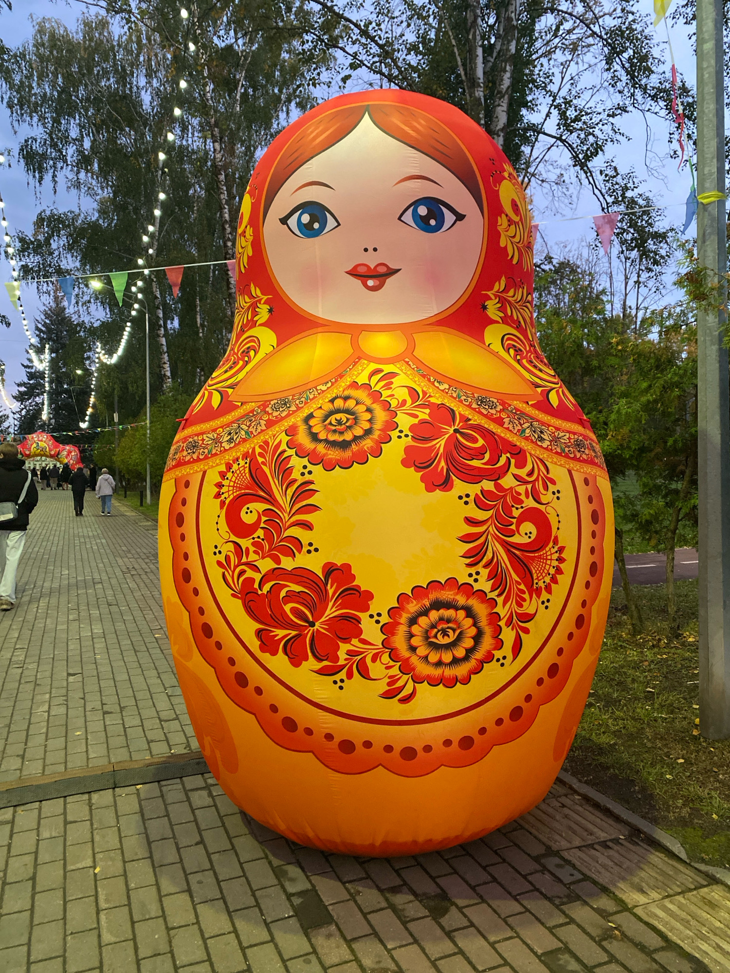 An inflatable Matryoshka doll, approximately 3 meters tall, stands on a paved walkway. The doll is predominantly yellow and orange, adorned with red floral patterns in the Khokhloma style. It has large blue eyes, red lips, and a brown hairline. The scene is outdoors, with trees lining the walkway and strings of lights and small triangular flags strung between them, suggesting a festive atmosphere. In the background, several people are walking along the path, and a road with sparse trees is visible to the right. The lighting indicates it is dusk. This installation is located in the city of Ivanteyevka, Russia, specifically on a pedestrian street.