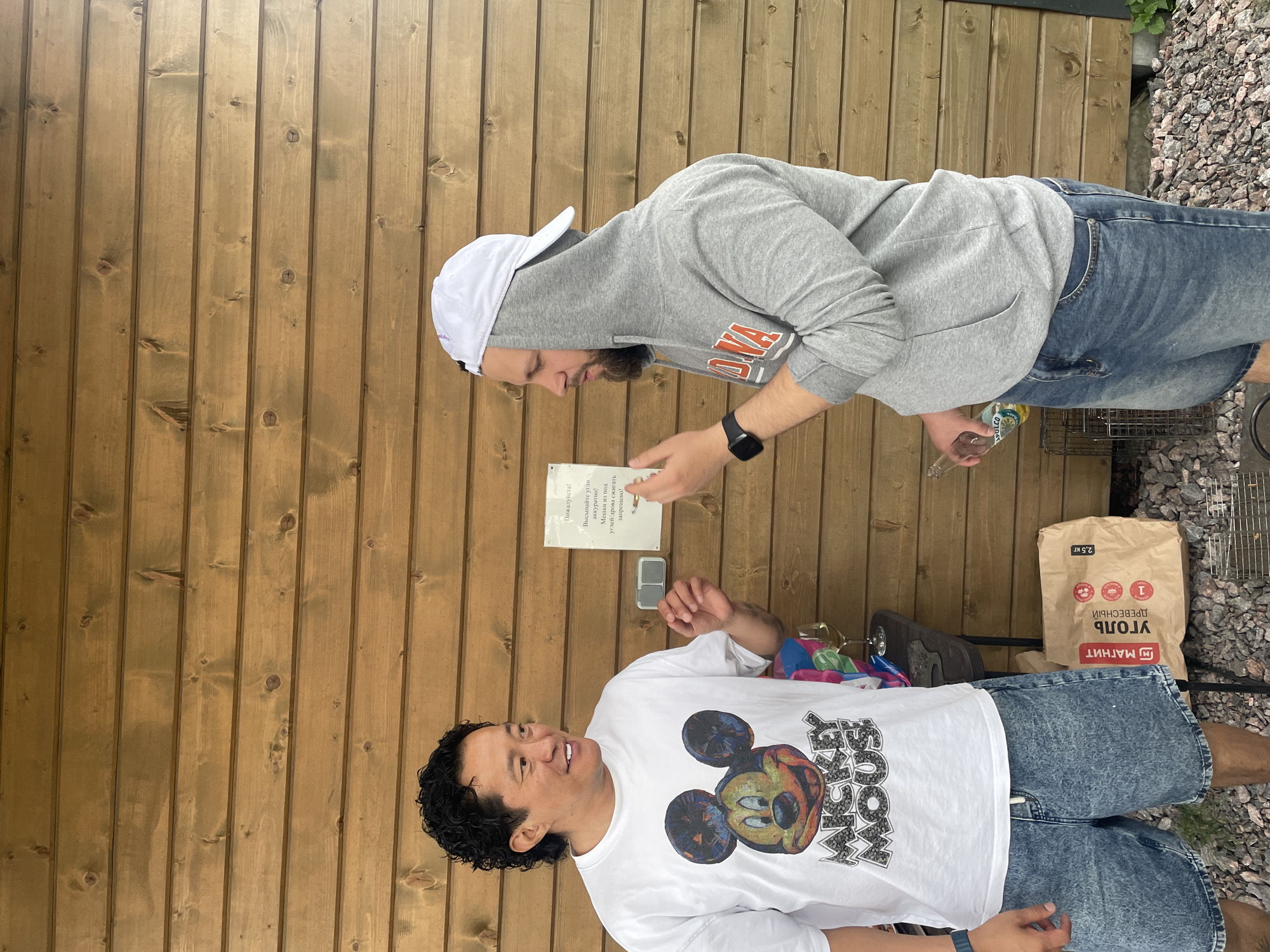 Two men are standing outdoors in front of a wooden wall. The man on the left is wearing a white t-shirt with a Mickey Mouse graphic and denim shorts. He has dark, curly hair and is smiling. The man on the right is wearing a grey hooded sweatshirt, blue denim shorts, and a white baseball cap. He has a beard and is looking at a sign on the wall. He is holding a glass bottle. A bag of charcoal is on the ground to the right of the man in the t-shirt, next to a grill. A small table with a glass of wine and other items is behind the man in the t-shirt. The scene appears to be in Novo-Tuutari, Russia, likely a residential area or a recreational space, possibly a dacha.