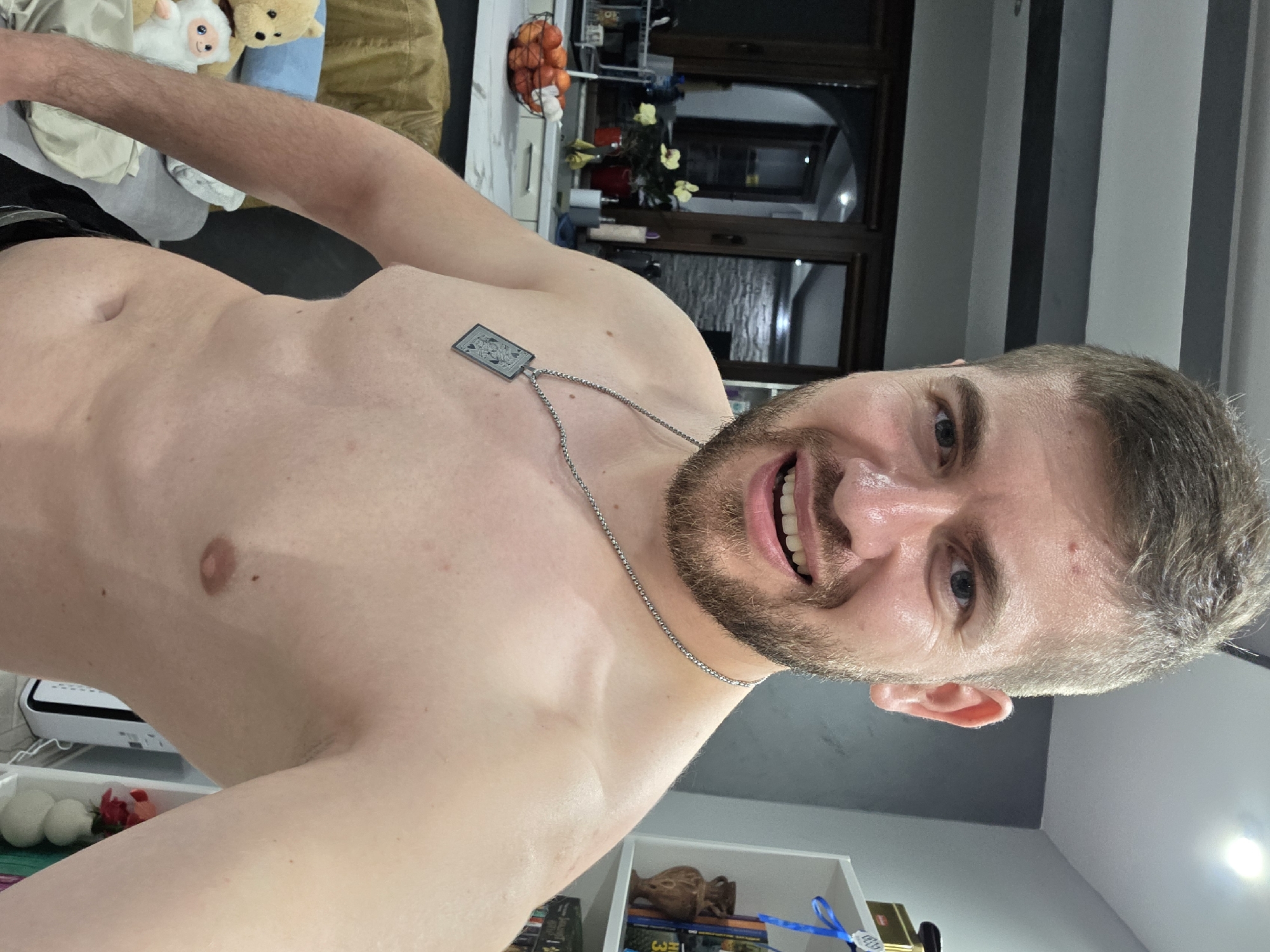 A close-up, low-angle shot of a Caucasian man with short, light-brown hair and a trimmed beard, wearing a silver chain necklace with a square pendant, shirtless and smiling towards the camera. The background shows a blurred interior of a room. There's a glimpse of furniture, decorations, and possibly a kitchen area with a fruit basket. The image does not provide any clues about the location of Yagodovo, Bulgaria.