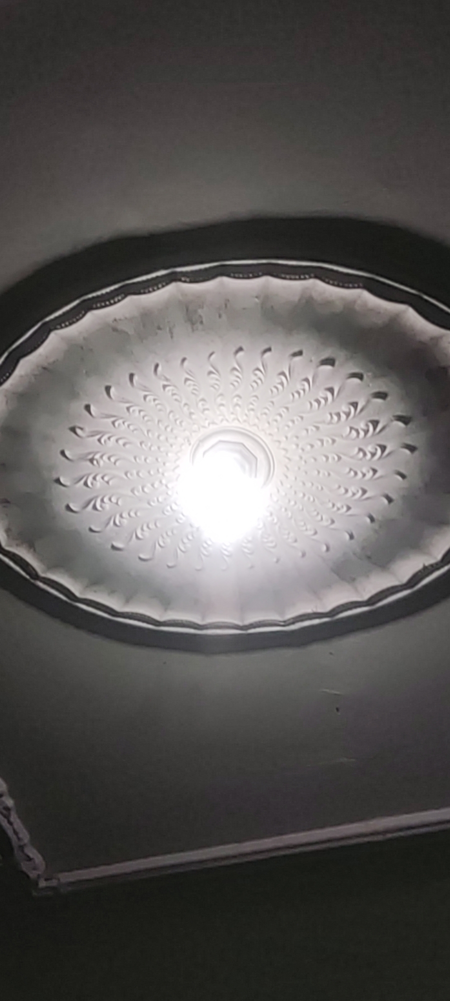 An overhead, medium-range shot shows a bright, white light emanating from a decorative ceiling fixture. The fixture has a radial, fan-like pattern with intricate detailing. The ceiling itself appears to be a neutral color, possibly white or light gray, with some subtle texture. The lower portion of the image shows a dark, indistinct area, possibly part of the room's walls or a shadow. The location is specified as Gebang, Indonesia.