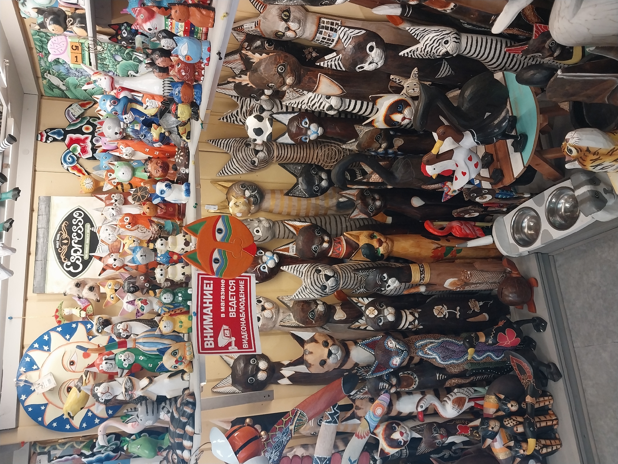 The image displays a vibrant and densely packed collection of handcrafted wooden animal figures, primarily stylized cats, in what appears to be a retail store or market stall in Sochi, Russia. The scene is dominated by numerous carved animals, with elongated bodies and various painted designs, arranged vertically against a light-colored wall.
The figures showcase a wide range of colors and patterns, from natural wood tones with painted stripes and spots to brightly painted examples in blue, green, orange, red, and white. Many of the cat carvings feature distinct facial expressions and intricate details, suggesting unique artisanal work. Other animals like zebras, a flamingo, and ducks are also visible among the cat figures.
In the background, a large, circular wall decoration with a sun and moon motif in blue, white, and orange is partially visible on the left. Above it, a rectangular sign reads "GREAT TASTE Espresso QUALITY BEANS." A prominent red and white sign, translated from Russian, states: "ATTENTION! Video surveillance is being conducted in the store." Towards the top right, a grey pet feeder with two metal bowls is visible. The overall impression is that of a bustling shop selling decorative and unique wooden handicrafts. The lighting is bright and even, typical of an indoor commercial setting.