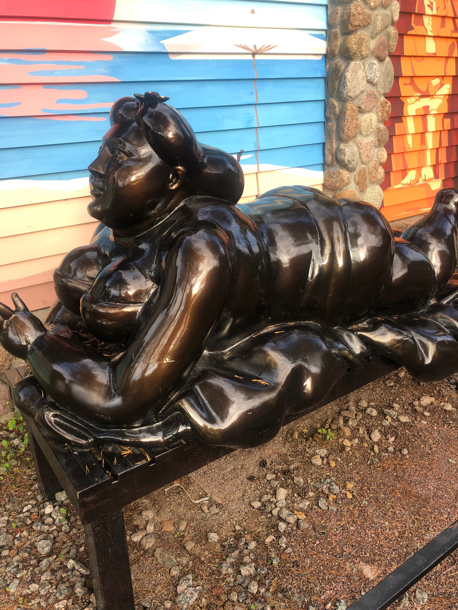 A dark, glossy bronze sculpture of a reclining, overweight figure lies on a dark wooden bench. The figure has exaggerated curves and appears to be on display outdoors. Behind the sculpture is a colorful, horizontally planked wall with a mural of abstract shapes and patterns in red, pink, and blue. To the right of the planked wall is a section of wall constructed of stacked, rough-hewn stones, followed by another planked section with an orange mural depicting stylized human figures. The ground around the bench is covered in gravel, small rocks, pine needles, and a few scattered pine cones. The scene suggests an outdoor art installation or decorative element in a public space. The location is identified as Интурист Снт, Russia.