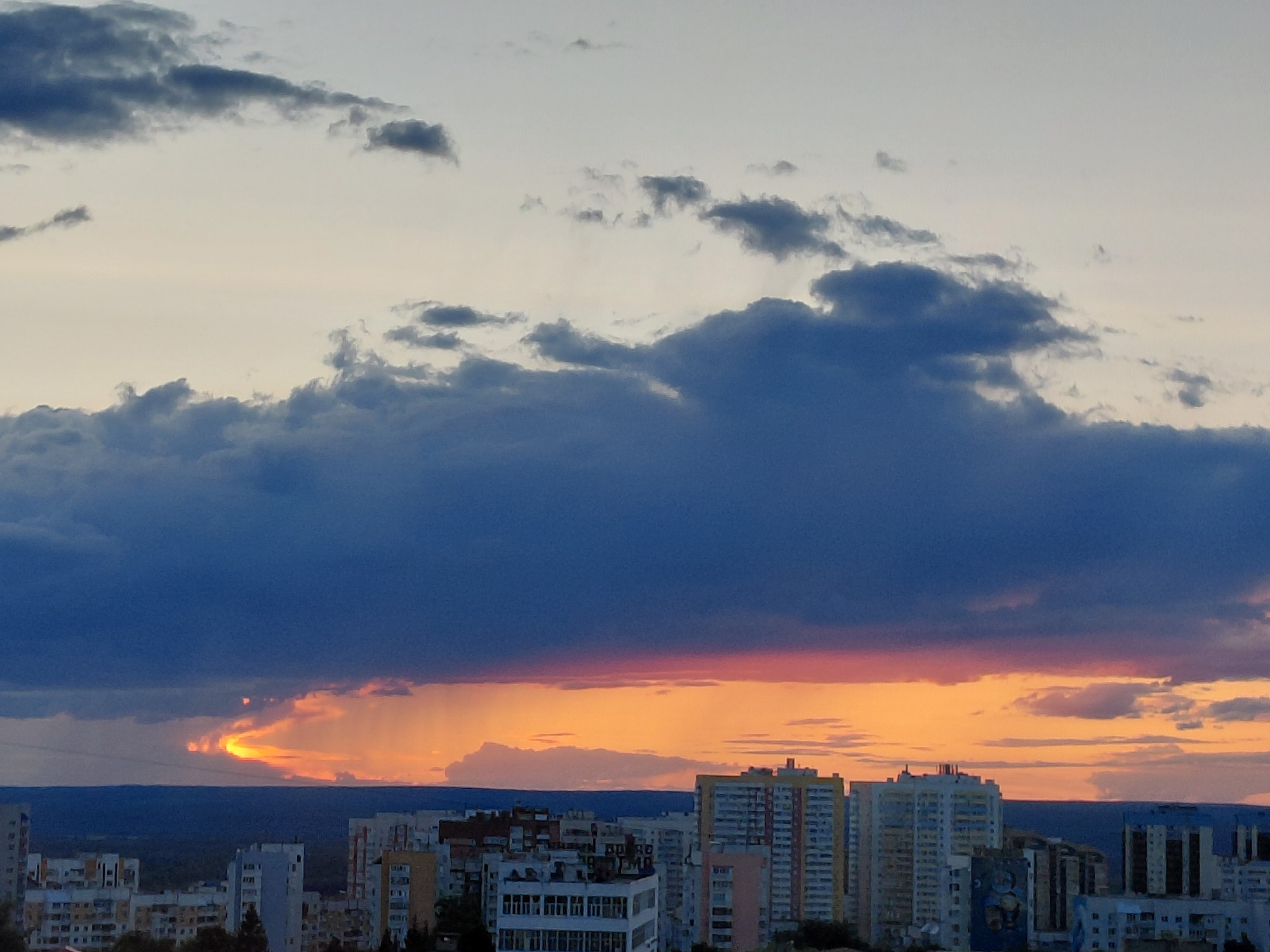 The image captures a dramatic sunset over the city of Novokuybyshevsk, Russia. A vast expanse of dark, brooding clouds dominates the upper portion of the frame, contrasting sharply with a band of vibrant orange and yellow light breaking through the horizon. This radiant glow illuminates a strip of sky, suggesting the sun is either setting or has just dipped below the visible horizon. Below the colorful sky, the silhouettes of numerous apartment buildings and other urban structures are visible, creating a dense cityscape. The buildings are varied in height and architectural style, indicative of a typical Soviet-era urban development. The overall atmosphere is one of natural beauty colliding with urban reality, with the intense colors of the sunset painting the sky above the densely populated city.