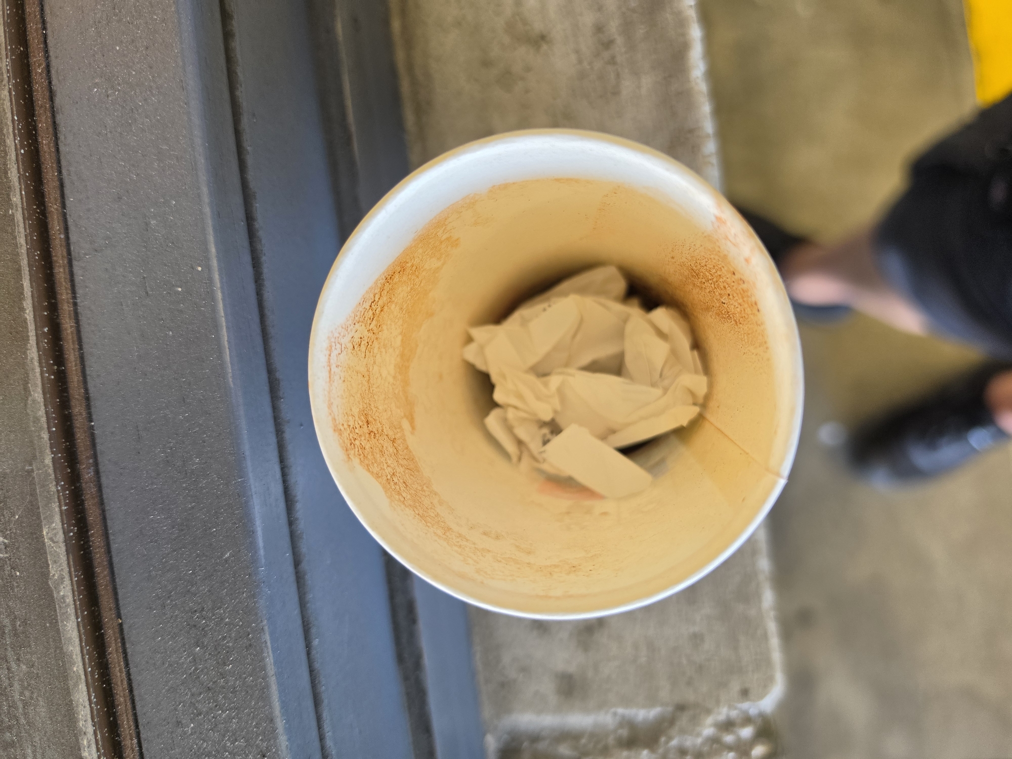 An overhead medium shot shows a white paper cup with crumpled paper inside, placed on a gray, textured surface. The rim of the cup has an orange-brown residue. In the background, a dark gray textured wall with horizontal blue accents is visible. The lower portion of the image shows a person's legs and feet, suggesting they are standing near the cup. The scene is devoid of any identifiable landmarks that would suggest a specific location within Frankfurt, Germany.
