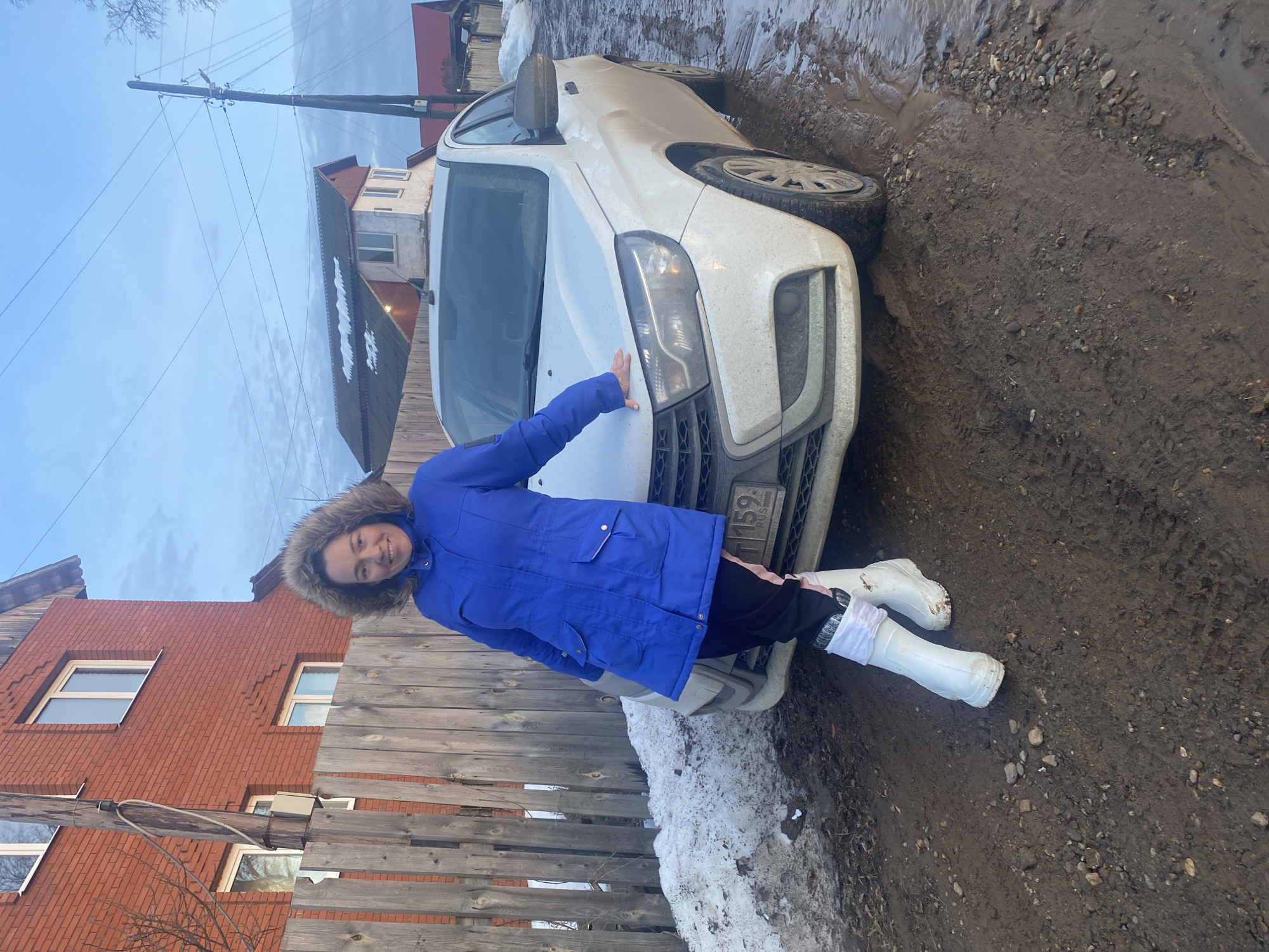 A woman stands posing on a muddy dirt road in Olkhovka, Russia. She is positioned slightly to the left of a dirty white sedan, resting her right hand on its hood. The woman, appearing to be an adult in her 30s-40s, wears a royal blue hooded winter coat with fur trim, black pants with a pink stripe, and white rubber boots with white fabric cuffs. She is smiling and looking directly at the camera.
The white sedan, likely a Lada, displays a partially visible Russian license plate with "159 RUS." Its front bumper and lower body are coated in mud. The ground is a heavily rutted and puddled dirt road, showing tire tracks and small rocks. Patches of dirty, melting snow are visible along the wooden fence to the left of the car.
In the background to the left, a two-story red brick house with white-framed windows is visible behind the wooden fence. To the right, a wooden utility pole with multiple overhead power lines stands against a pale blue, partly cloudy sky. Further in the background, another house with a grey roof and a lighter facade, along with a red-roofed structure, can be seen. The scene suggests a late winter or early spring setting.