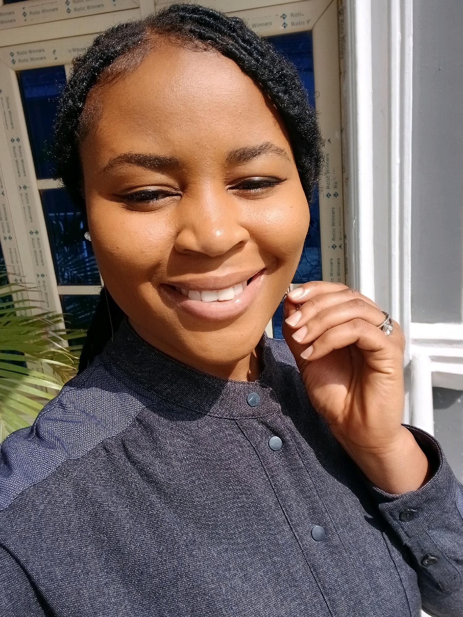 A headshot shows a woman with dark skin and braided hair smiling in front of a window. The woman is wearing a dark blue collared shirt with button details. She is wearing a ring on her left ring finger. The window frame has writing on it. The background is out of focus. The lighting suggests the photo was taken during the day outdoors. The woman is likely at her residence or a place with a similar window in Gura Top, Nigeria.