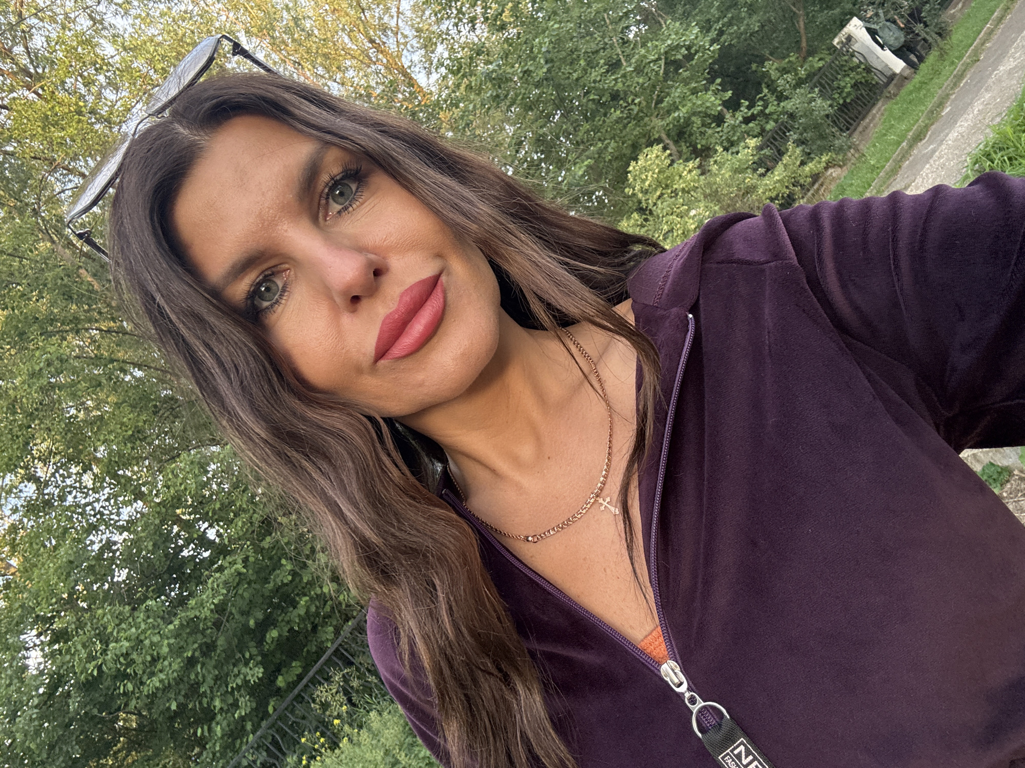 A woman is taking a selfie outdoors in Chaikovsky, Russia. She has long, wavy brown hair and is wearing a dark purple velour zip-up hoodie over an orange top. A gold necklace with a cross pendant is visible around her neck. She is also wearing sunglasses perched on her head. The background consists of lush green trees and a sidewalk.