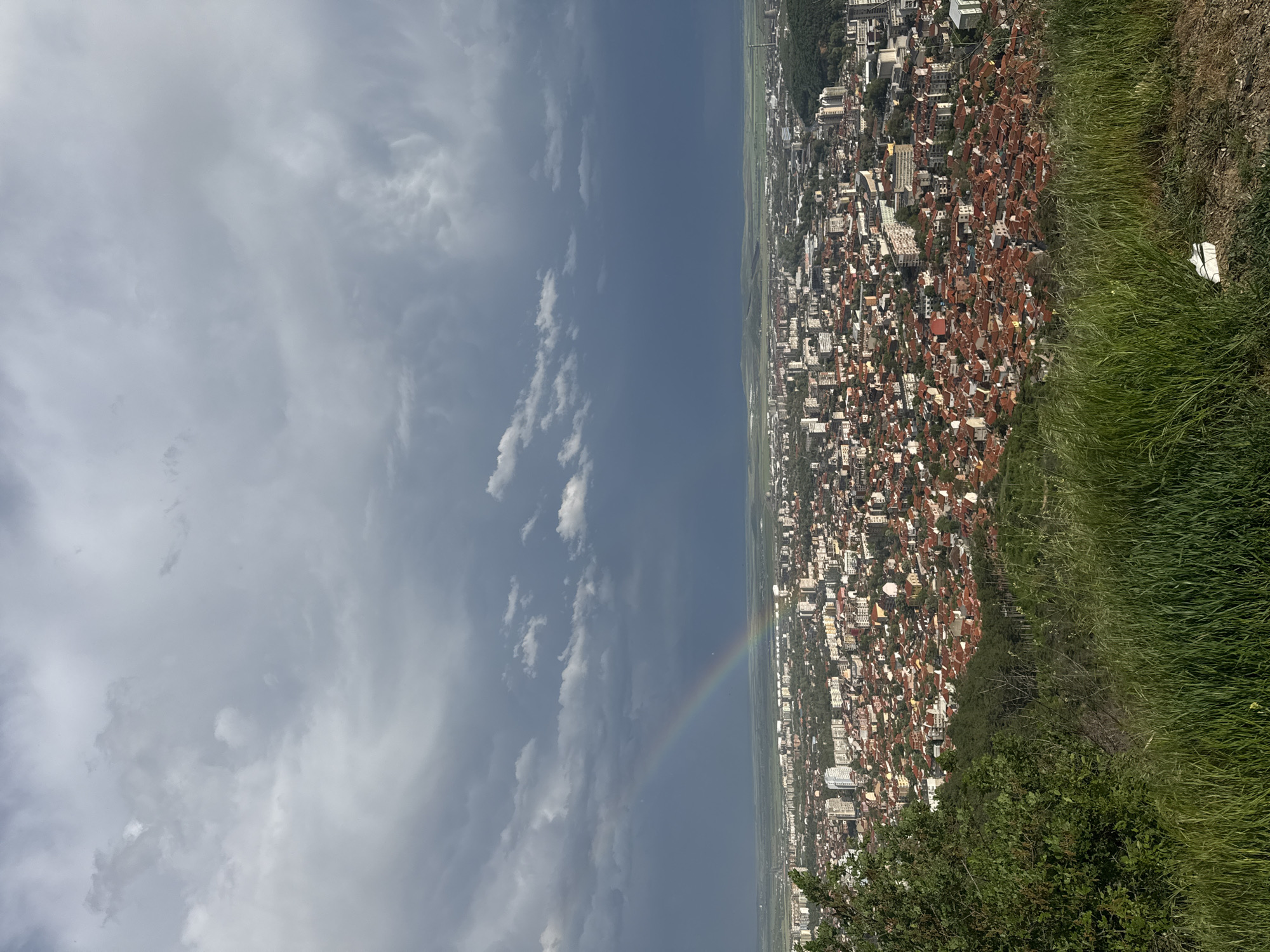 The image shows a city viewed from an elevated position, possibly a hillside or high-rise building. The city, identified as "Unknown, Unknown," is densely built with a mix of residential and commercial structures. Many buildings have reddish roofs. Green grass and vegetation appear in the lower right of the frame, indicating the vantage point is at the edge of a natural area. The sky is predominantly cloudy, with a section of blue sky visible. A faint rainbow spans across the cityscape.