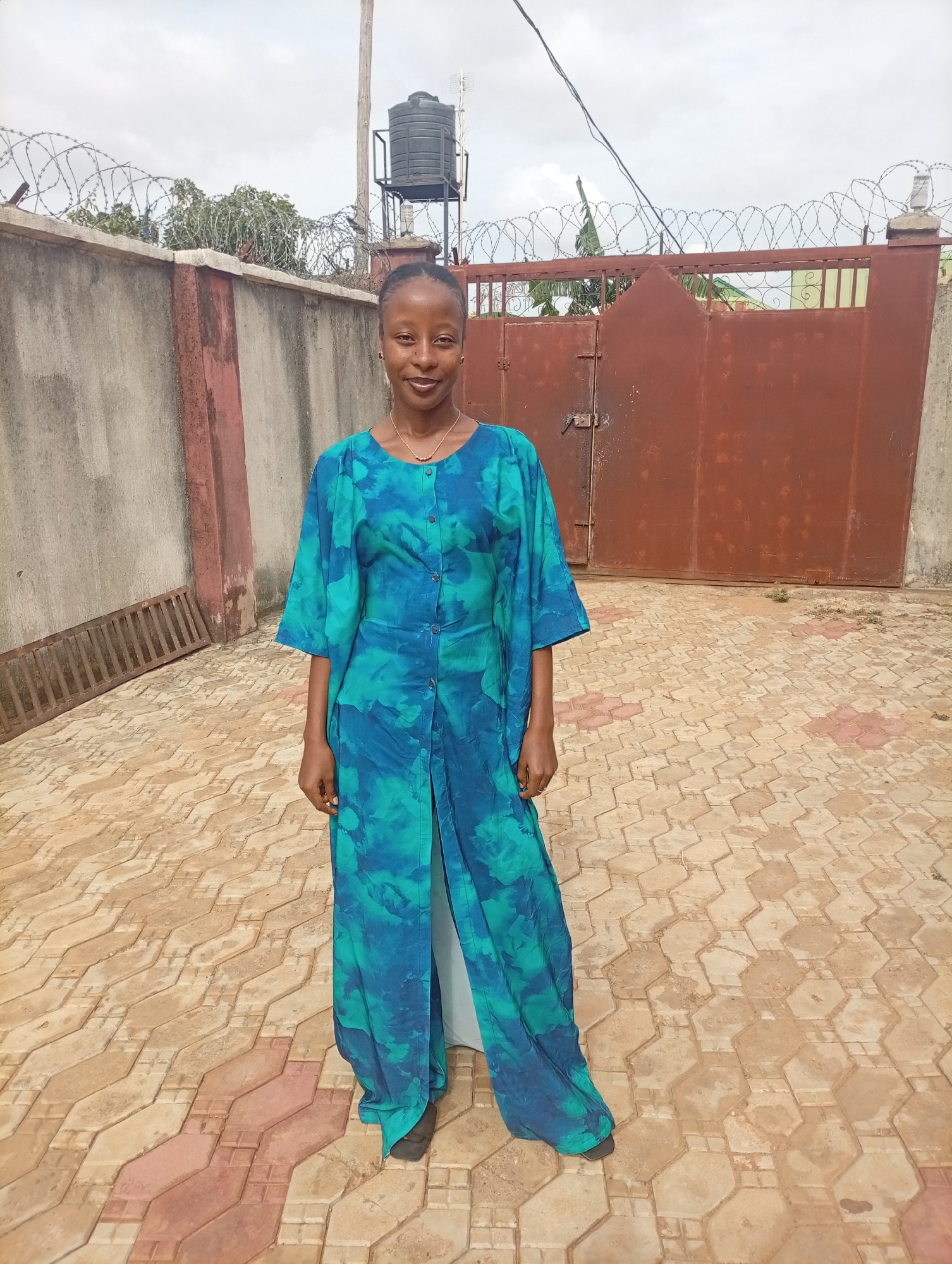 A woman stands in a courtyard with a patterned brick surface in Rido, Nigeria. She wears a blue and turquoise tie-dye-patterned outfit consisting of a long-sleeved tunic-style top and long pants. Her hair is pulled back, and she wears a gold necklace.
Behind her is a rusted, orange-brown gate, set into a concrete wall topped with barbed wire. To her left, is another section of concrete wall, and an object resembling a metal grate is leaning against it. A black water tank is mounted on a metal support structure in the background. Above, the sky is overcast. Overhead power lines are visible.