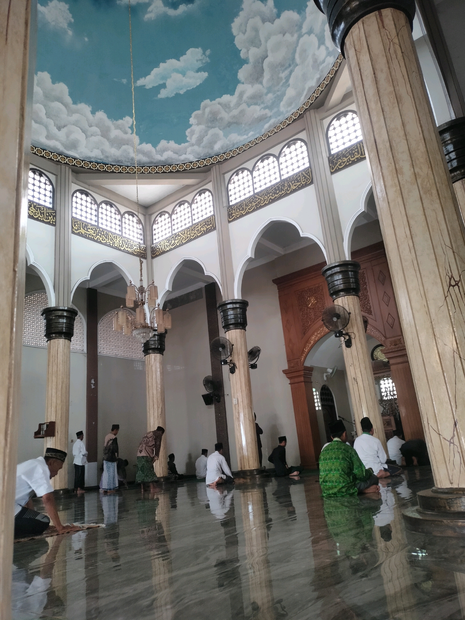The image captures the interior of a mosque in Kutosari, Indonesia, characterized by a domed ceiling painted with clouds and a blue sky. Large, fluted columns with dark capitals line the space, supporting the structure. Arched windows with intricate latticework are visible along the upper walls, above decorative Arabic calligraphy. A chandelier hangs from the ceiling, illuminating the polished, reflective floor. Several men are present, some seated on the floor, while others are in various prayer positions, suggesting ongoing or recently completed religious observances. The atmosphere appears serene and spiritual.