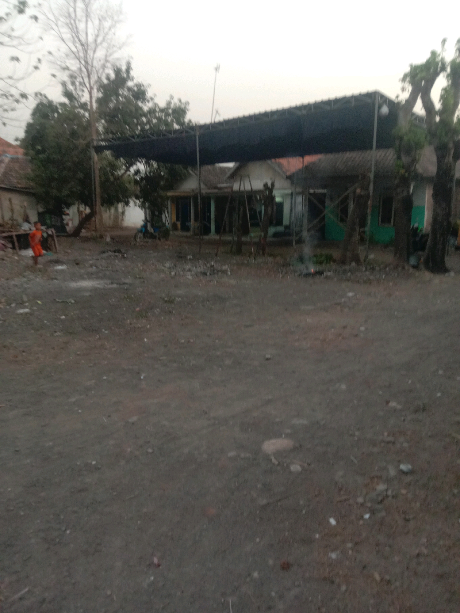 In Karang Anyar, Indonesia, a rural area is depicted with a dirt ground and a few structures. A small child, wearing an orange shirt and shorts, stands near a wooden structure on the left side of the image. Behind the child, a dirt road leads to several buildings with dark roofs and walls. A motorcycle is parked in front of one of the buildings. In the center of the image, a large, dark canopy stretches over an open area, supported by poles. A small fire is burning near the base of a tree under the canopy, emitting a thin plume of smoke. Trees with sparse foliage are scattered throughout the scene, and the sky is overcast.