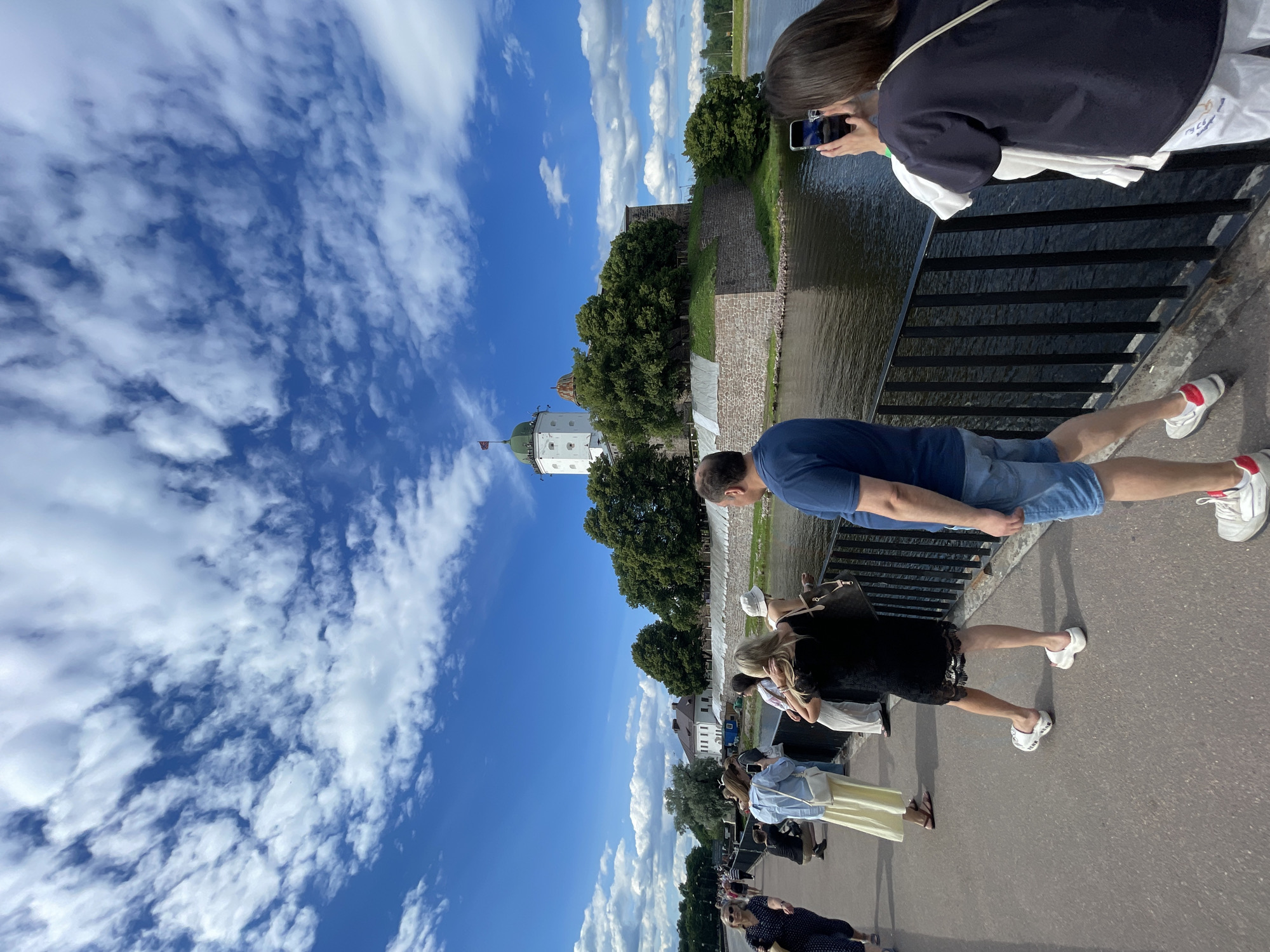 The image shows a view from a bridge in Vyborg, Russia, looking towards Vyborg Castle. The sky is blue with white, wispy clouds. Several people are walking on the bridge. In the foreground, on the right, a person in a dark blue shirt and denim shorts is facing away from the camera. To their left, a woman with dark hair is holding up a smartphone to take a picture. In the middle ground, a woman in a black dress is walking away from the camera, and further to the left, a woman in a long yellow skirt and a light blue top is walking towards the camera, holding a phone. Behind the people, a black metal railing lines the edge of the bridge. The castle, a white tower with a green dome and a flag on top, sits on an island surrounded by water and trees. The castle has stone walls and is a prominent landmark.