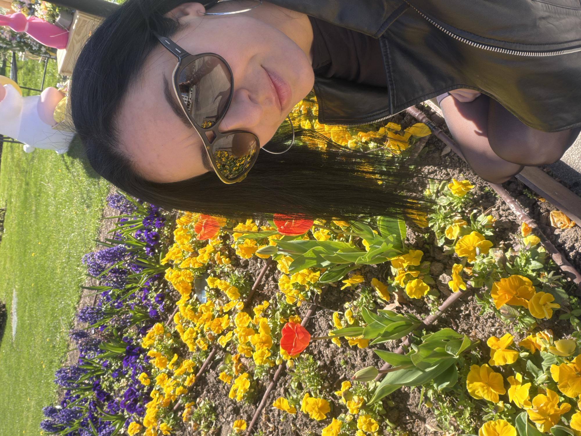 A woman with dark hair and sunglasses poses in a garden. She is wearing a black leather jacket and dark hosiery. In the background is a flower bed containing yellow and purple flowers, with red tulips interspersed. There is a grass lawn and a decorated animal figure visible in the background. The location is Pleven, Bulgaria.