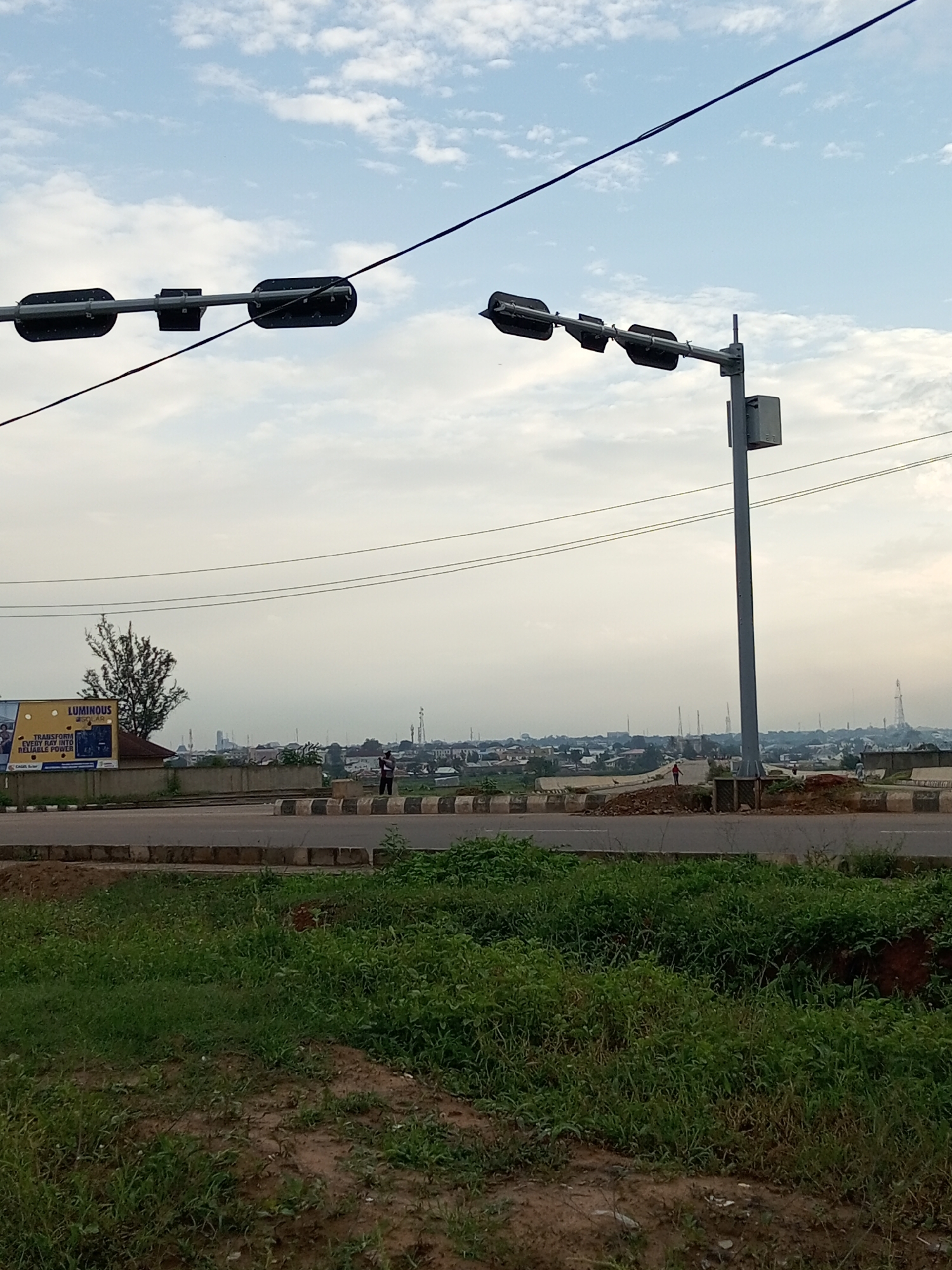 The image depicts an outdoor scene in Barden Yana, Nigeria. In the foreground, there is a patch of overgrown green grass and bare earth. A paved road runs across the middle ground, bordered by a concrete barrier with black and white stripes. Beyond the road, a distant city skyline is visible under a cloudy sky, with numerous telecommunication towers scattered across the landscape.
Two traffic light structures are prominent in the upper portion of the image. The first, on the left, is a horizontal bar with two black signal heads. The second, on the right, is mounted on a tall grey pole and also features two black signal heads. A grey electrical box is attached to the pole of the second traffic light.
Several individuals are present in the mid-ground. One person, wearing dark clothing, stands near the road, and another, in a red shirt, is further back on the roadside. A large billboard with the text "LUMINOUS SOLAR" and "TRANSFORM EVERY RAY INTO RELIABLE POWER" is visible on the left side of the frame, partially obscured by a tree.
The time of day appears to be either early morning or late afternoon, given the soft lighting and overcast sky. The weather seems to be slightly hazy. The scene suggests an urban or semi-urban environment with ongoing development or infrastructure.