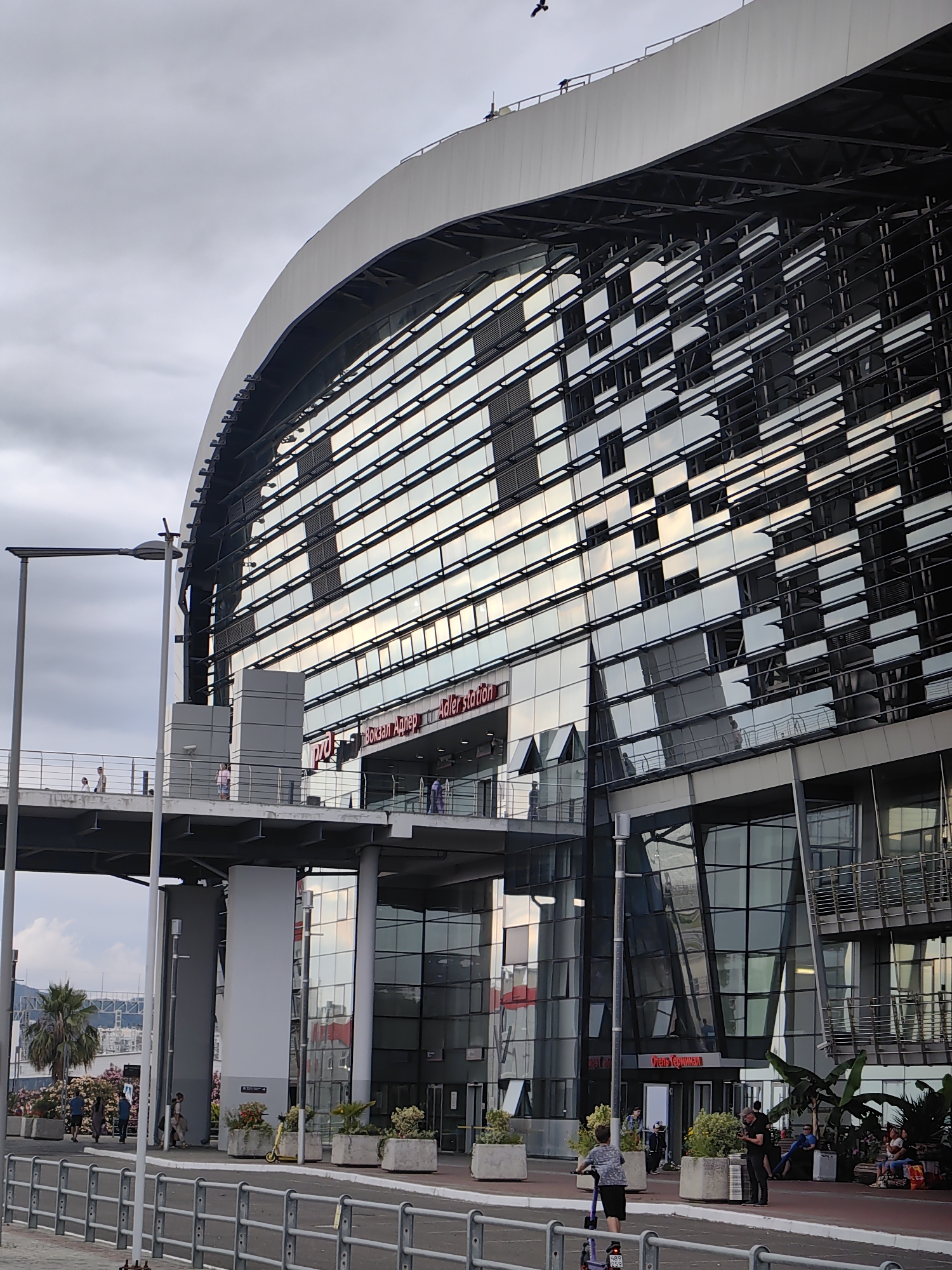 The image shows the Adler railway station in Sochi, Russia, on a cloudy day. The station is a modern building with a curved, glass facade reflecting the sky. A pedestrian walkway or bridge extends from the left side of the frame, leading to the station entrance. Several people are visible on the walkway and in front of the station. There are also potted plants and a yellow scooter on the ground level. The architecture of the station is prominent, with its sleek lines and extensive use of glass.