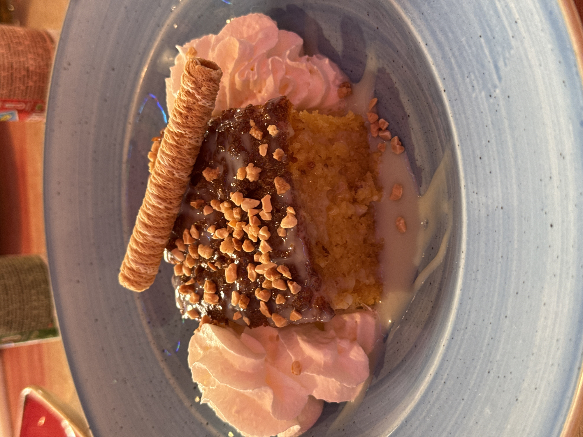 A top-down shot shows a dessert on a light blue plate. The dessert appears to be a slice of cake, potentially tres leches, drizzled with a light-colored sauce. A portion of the cake is glazed with a darker topping and sprinkled with chopped nuts. Two dollops of white whipped cream sit on either side of the cake slice. A cylindrical wafer stick lies alongside the cake. The setting suggests an indoor dining environment, potentially a restaurant. Based on the type of dessert and typical regional cuisine, the location could potentially be a restaurant in Madrid, Spain, given the popular dishes served there.